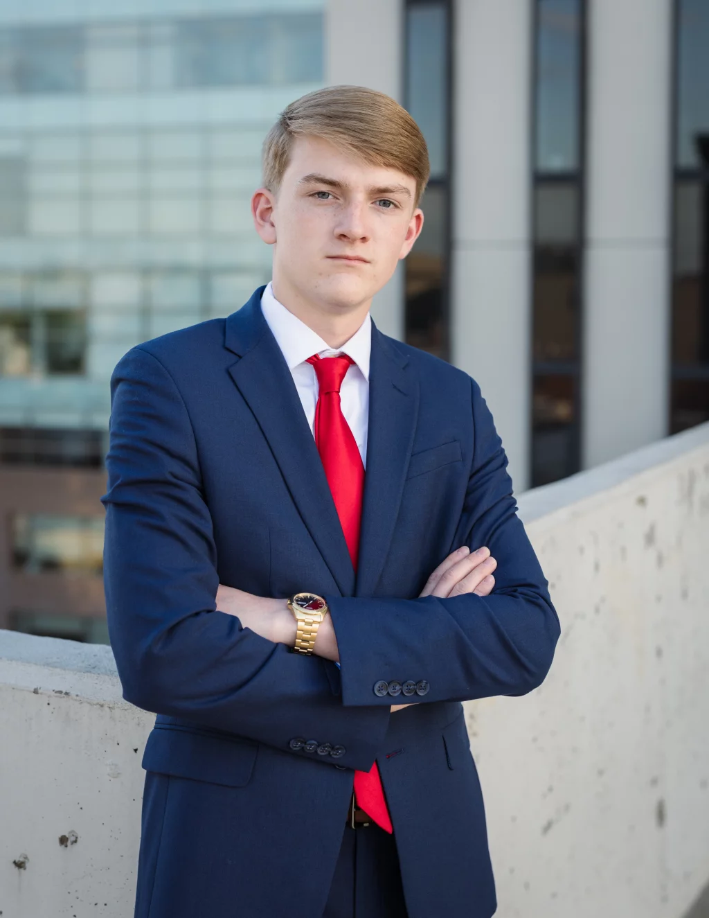 Macon's senior photos taken at Country Square and Downtown Greenville: Young man in a blue suit with a red tie and gold watch, standing with arms crossed in front of a concrete barrier and modern office building.