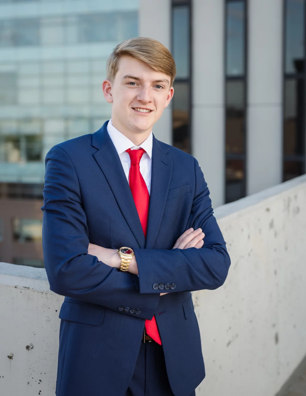 Macon's senior photos taken at Country Square and Downtown Greenville: Young man in a blue suit and red tie standing with arms crossed, smiling, outdoors against a backdrop of modern glass and concrete buildings.