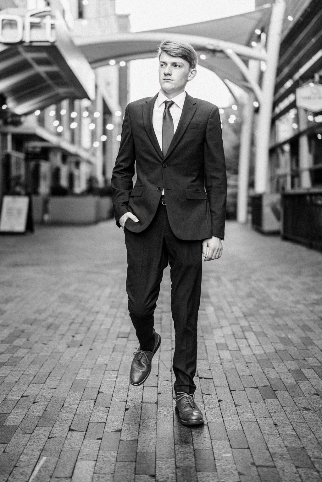 Macon's senior photos taken at Country Square and Downtown Greenville: Black and white photo of a young man in a suit walking confidently on a brick-paved urban street, with modern architecture and string lights in the background.