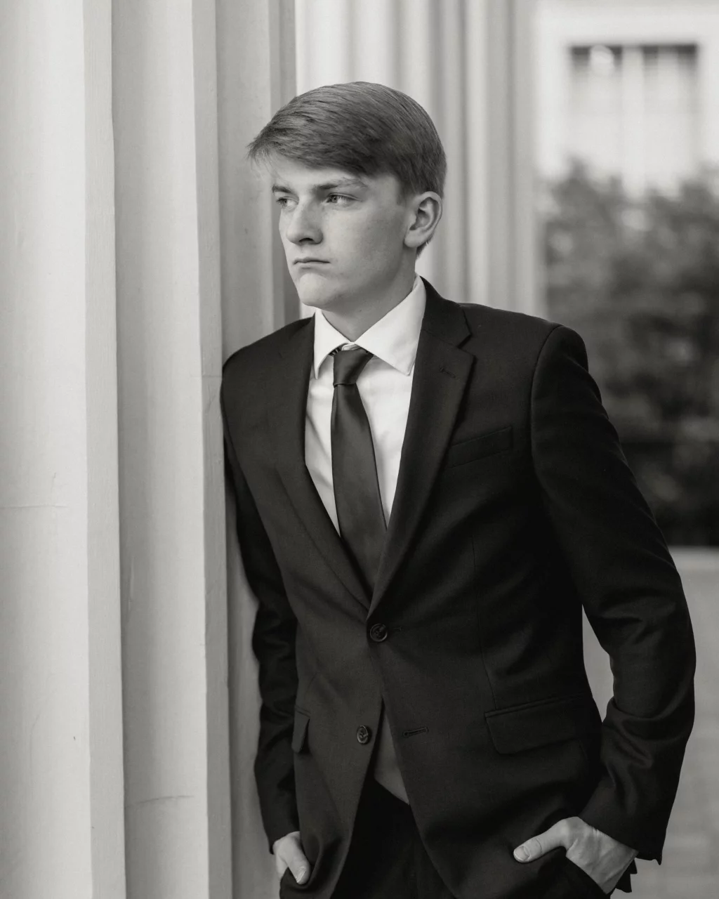 Macon's senior photos taken at Country Square and Downtown Greenville: Black and white photo of a young man in a suit and tie, standing with hands in pockets, leaning against a column and looking to the side with a serious expression.