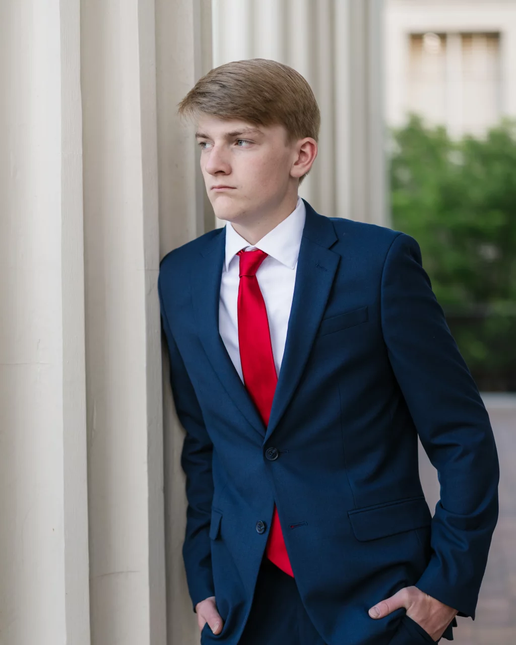 Macon's senior photos taken at Country Square and Downtown Greenville: Young man in a blue suit and red tie stands with hands in pockets, leaning against a white column and looking to the side.