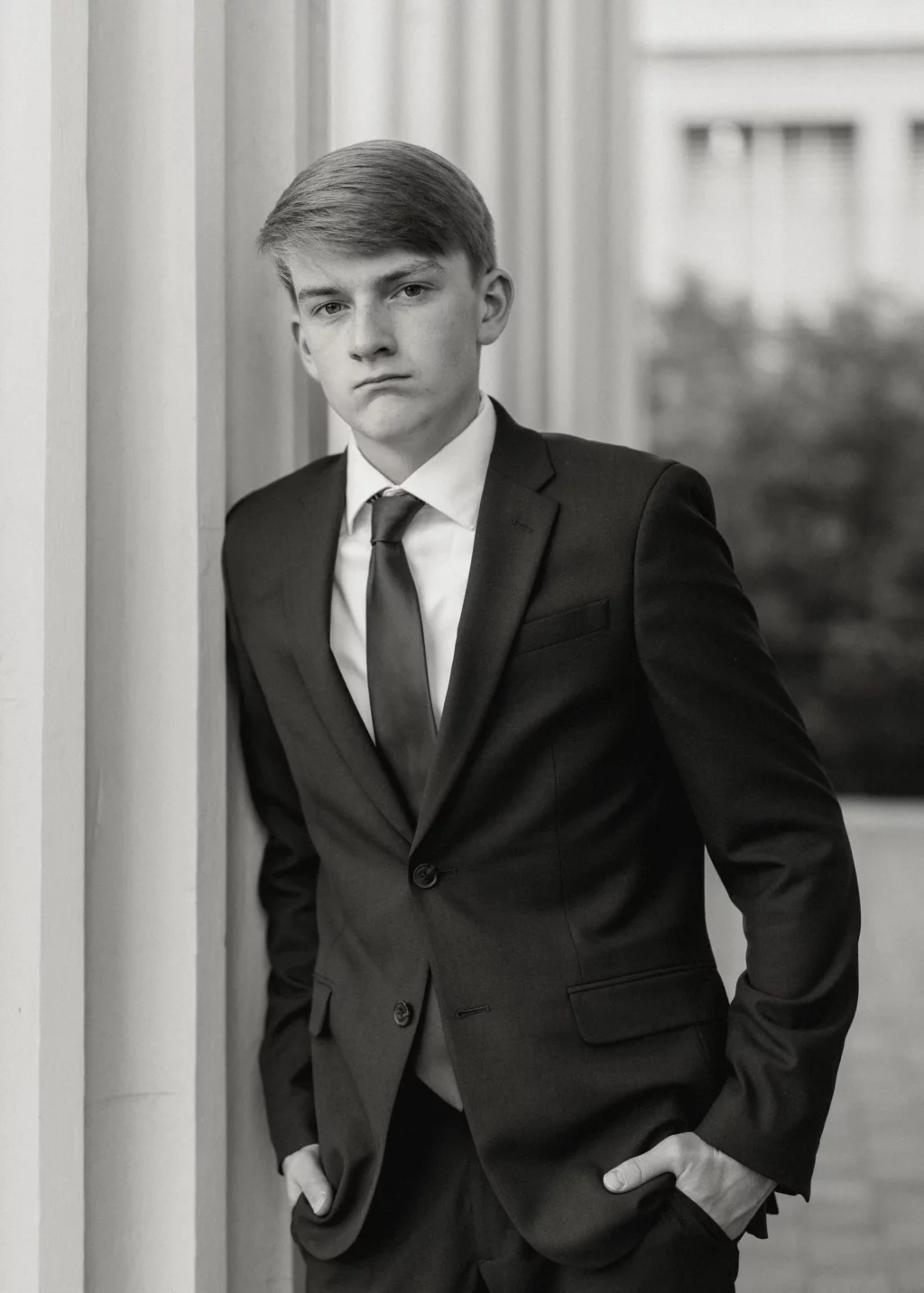 Macon's senior photos taken at Country Square and Downtown Greenville: Black and white portrait of a young man in a suit and tie, standing with hands in pockets and leaning against a column, looking seriously at the camera.