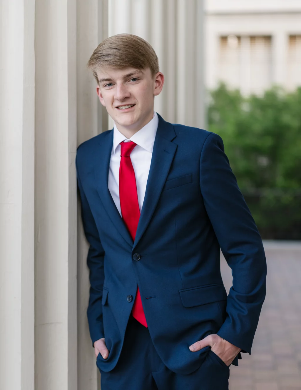 Macon's senior photos taken at Country Square and Downtown Greenville: Young man in a blue suit and red tie stands outdoors, leaning against a white column, with hands in his pockets and greenery blurred in the background.