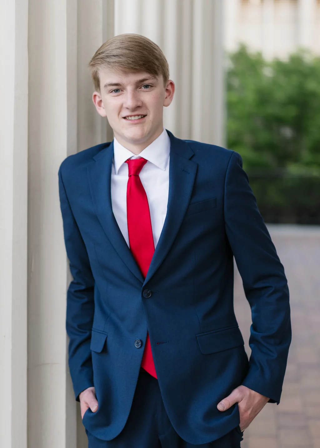 Macon's senior photos taken at Country Square and Downtown Greenville: Young man in a blue suit with a white shirt and red tie, standing outdoors against a column and smiling at the camera.