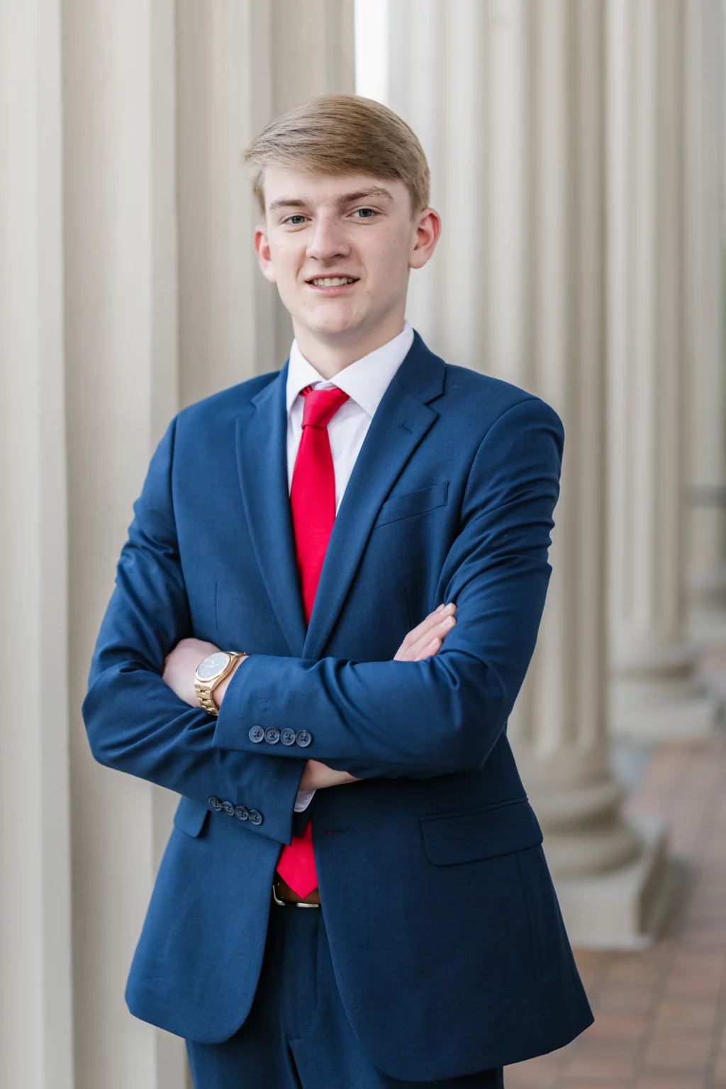 Macon's senior photos taken at Country Square and Downtown Greenville: Young man in a blue suit and red tie standing with arms crossed in front of tall, white columns.