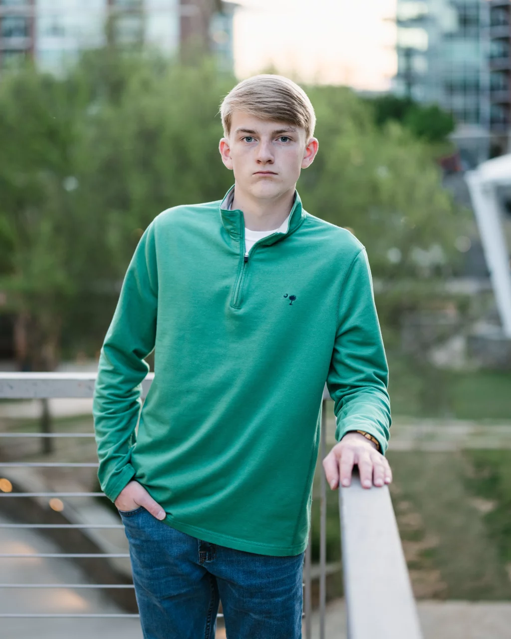 Macon's senior photos taken at Country Square and Downtown Greenville: Teenage boy with blond hair wearing a green pullover and blue jeans, standing outdoors and leaning on a railing with a neutral expression. Blurred trees and buildings are in the background.