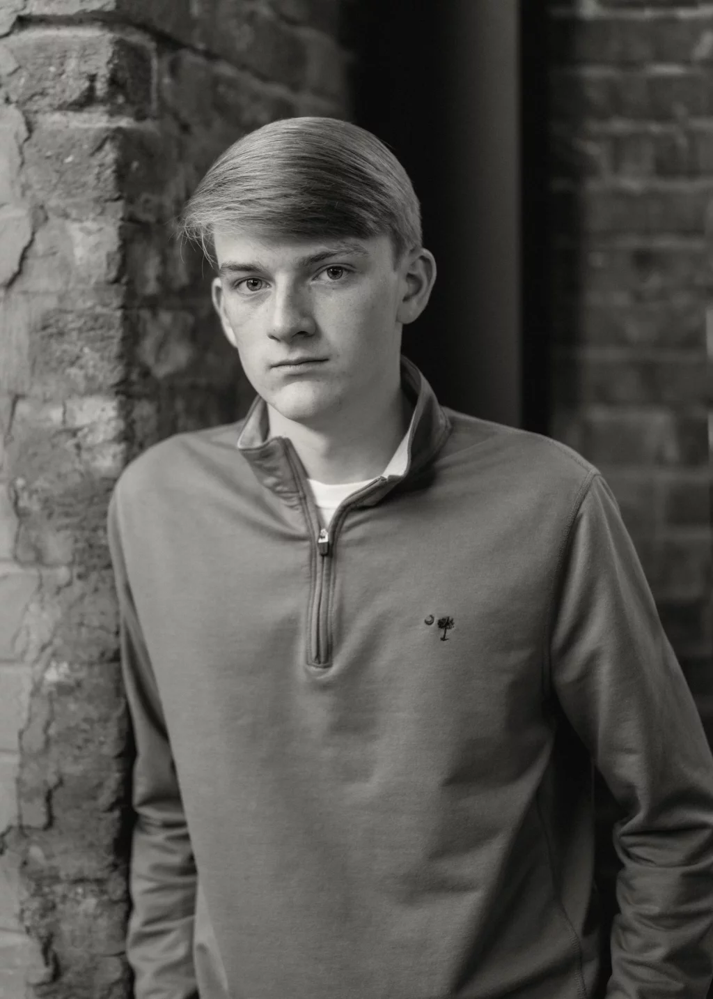 Macon's senior photos taken at Country Square and Downtown Greenville: Black and white portrait of a young person with short, light hair, wearing a quarter-zip pullover, standing against a rough brick wall and looking directly at the camera with a neutral expression.