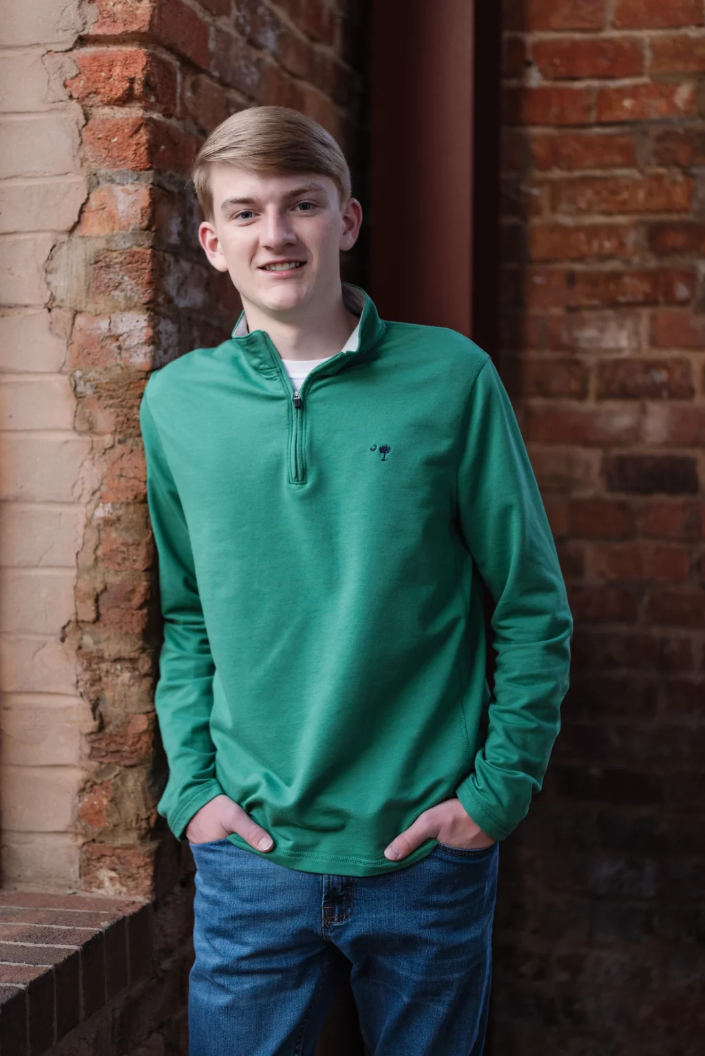 Macon's senior photos taken at Country Square and Downtown Greenville: Young person with short blond hair wearing a green pullover and blue jeans, standing and smiling with hands in pockets against a brick wall background.