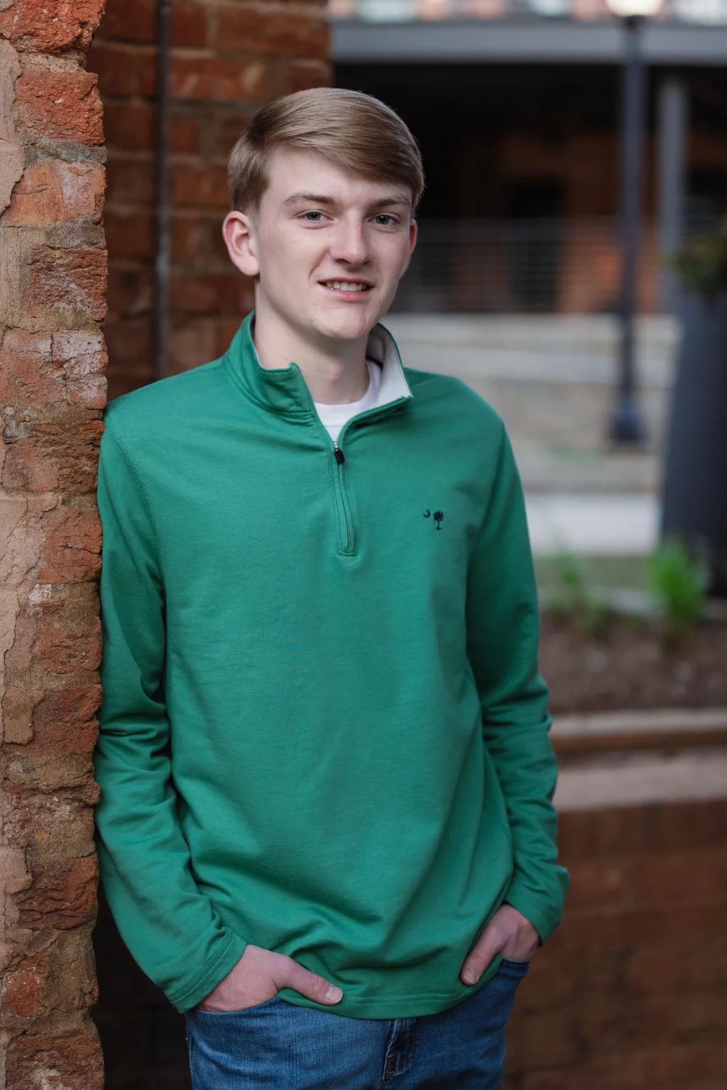 Macon's senior photos taken at Country Square and Downtown Greenville: Teenage boy with short light brown hair wearing a green quarter-zip pullover and blue jeans, standing with hands in pockets and leaning against a brick wall outdoors.