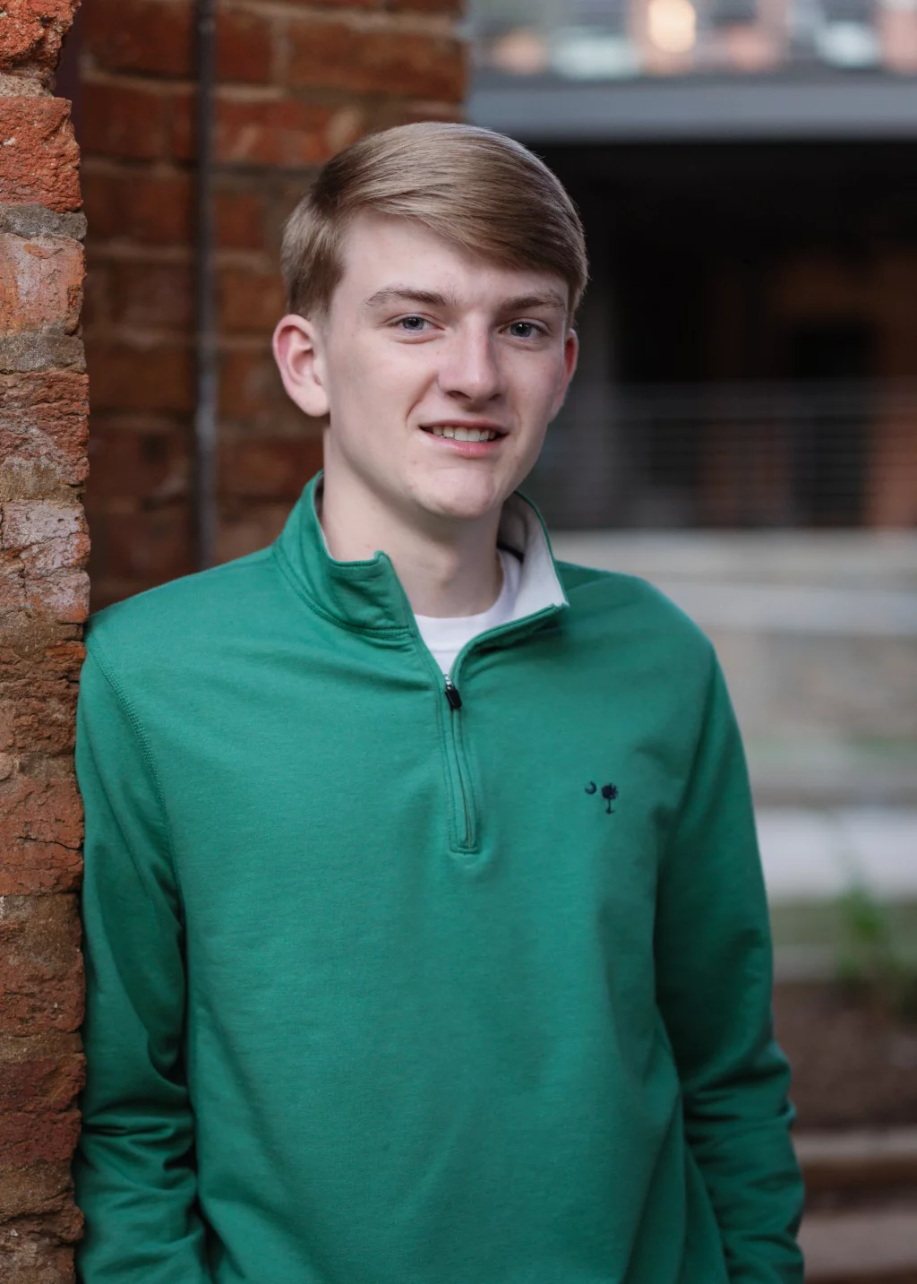 Macon's senior photos taken at Country Square and Downtown Greenville: Young man with short light brown hair wearing a green quarter-zip pullover, standing next to a brick wall and smiling at the camera.