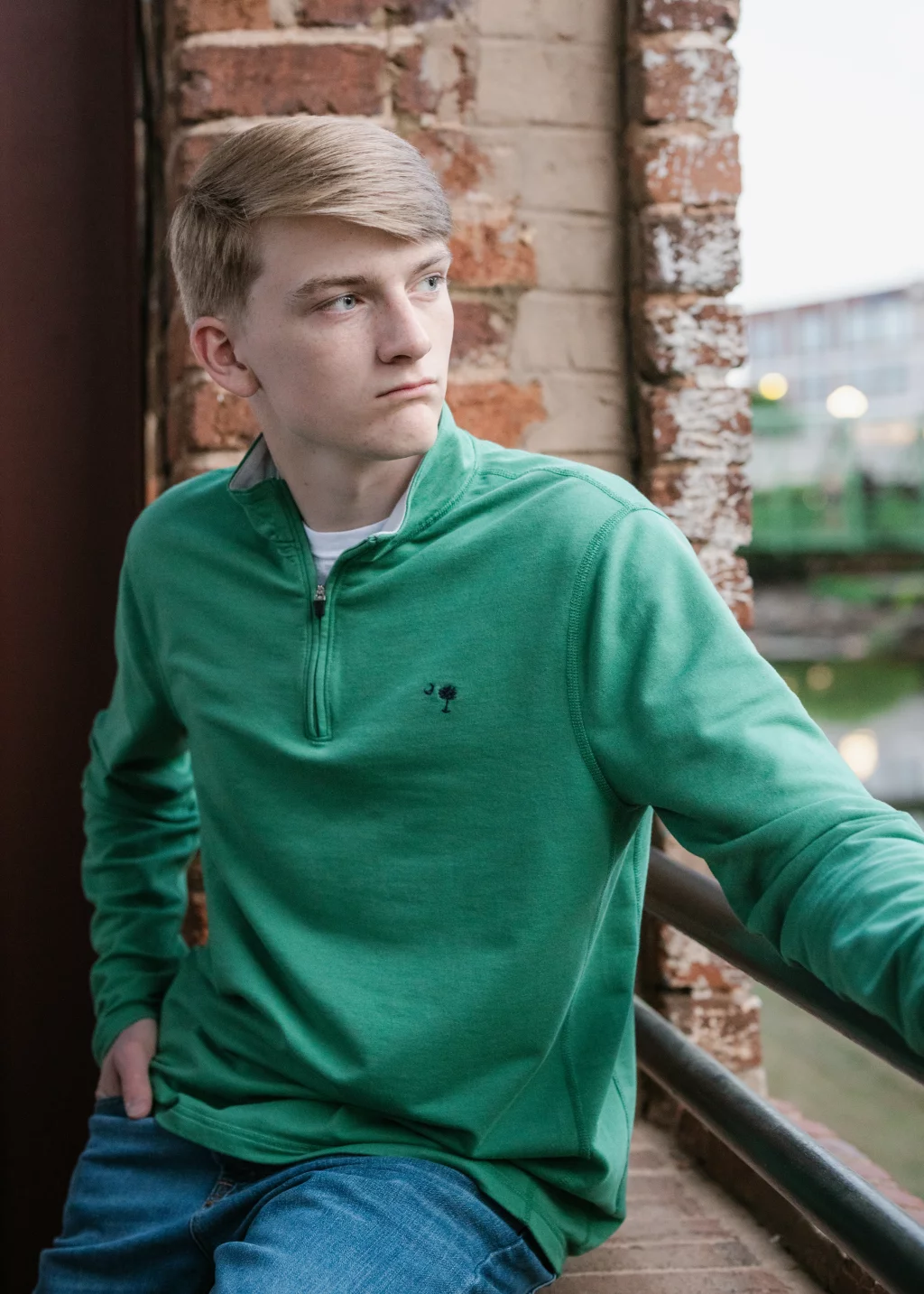 Macon's senior photos taken at Country Square and Downtown Greenville: Young man with short blond hair wearing a green quarter-zip pullover sits on a brick window ledge, looking to the side with a thoughtful expression.