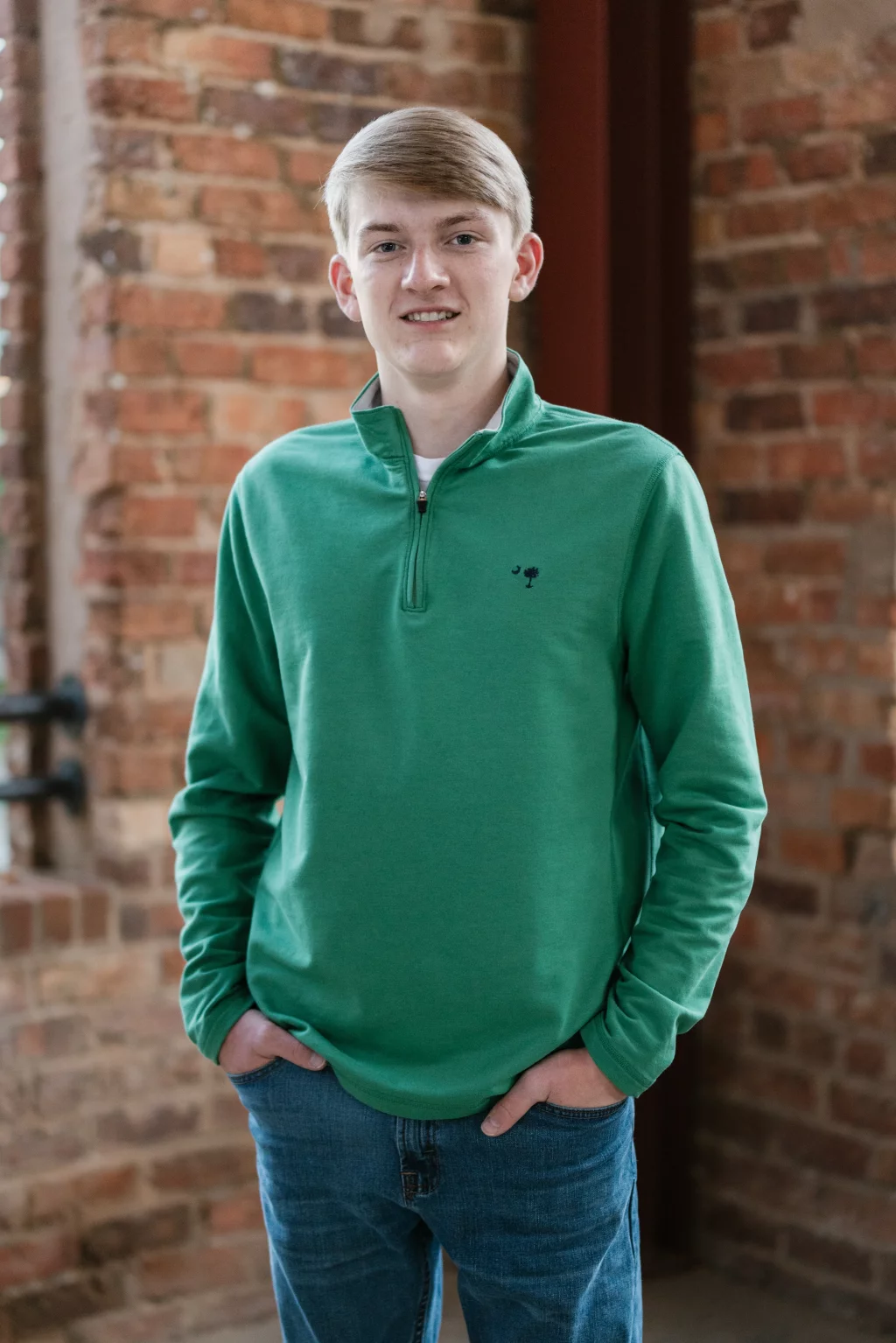 Macon's senior photos taken at Country Square and Downtown Greenville: Young man with short blond hair wearing a green quarter-zip pullover and blue jeans, standing with hands in pockets in front of a brick wall.
