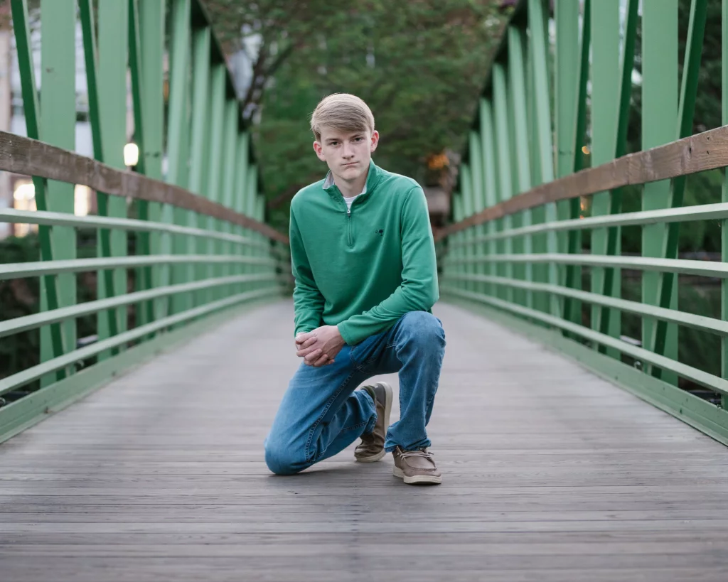 Macon's senior photos taken at Country Square and Downtown Greenville: Teenage boy in a green pullover and blue jeans kneeling on one knee in the center of a green metal pedestrian bridge, with trees in the background.
