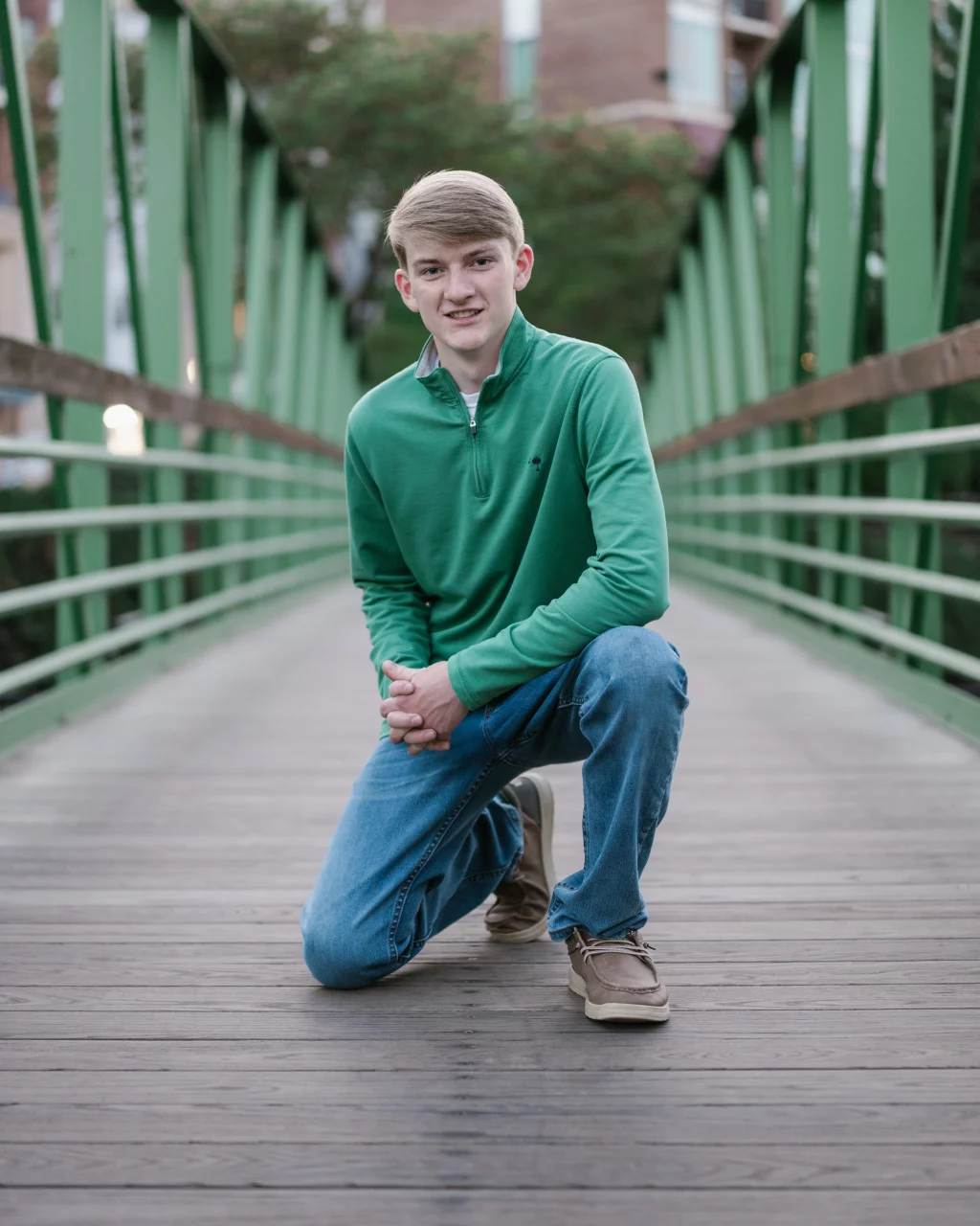 Macon's senior photos taken at Country Square and Downtown Greenville: Teen boy in a green pullover and jeans kneeling on a wooden footbridge with green railings, outdoors.