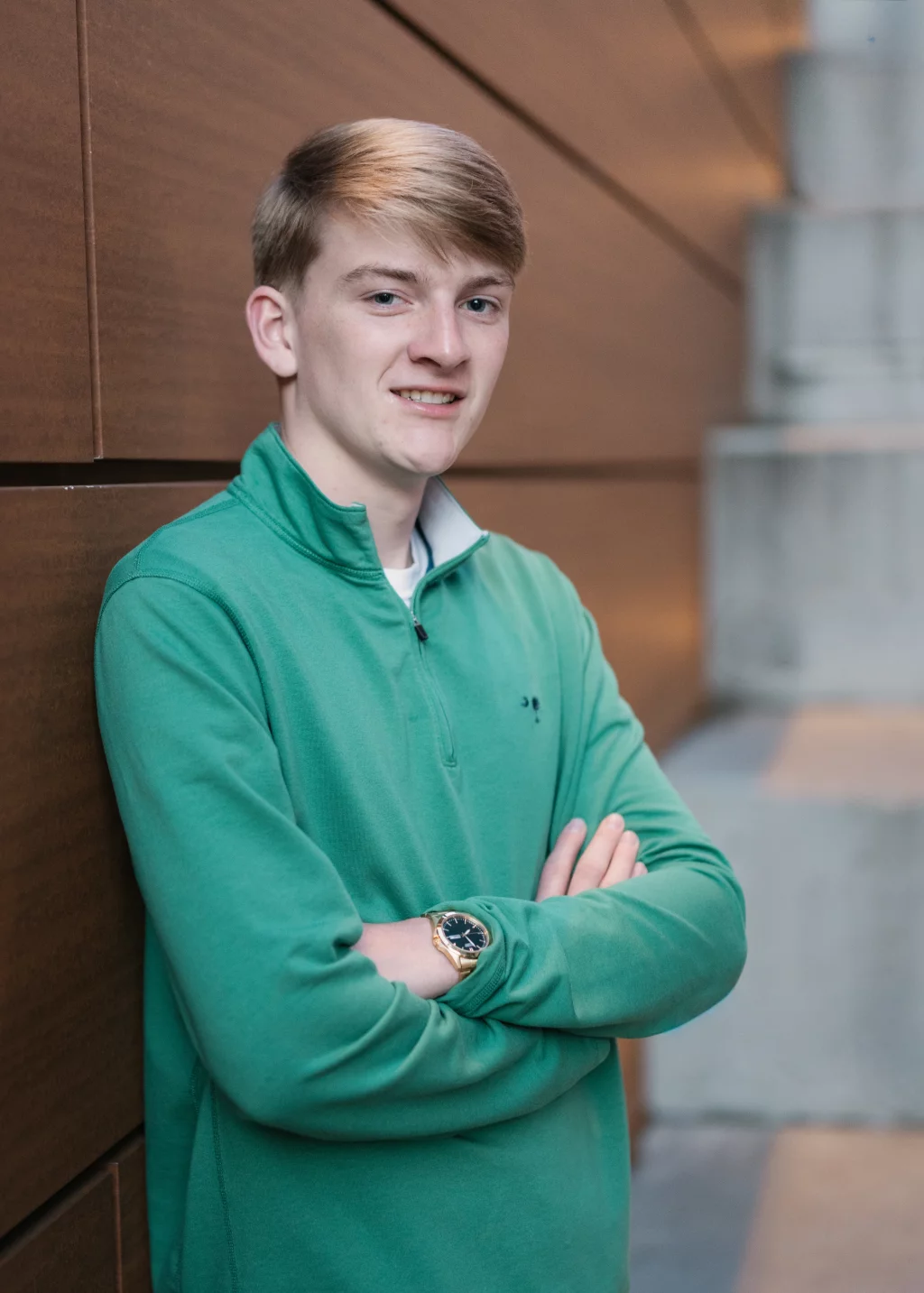 Macon's senior photos taken at Country Square and Downtown Greenville: Young person with light hair wearing a green pullover stands with arms crossed, leaning against a wood-paneled wall. The background features concrete steps.