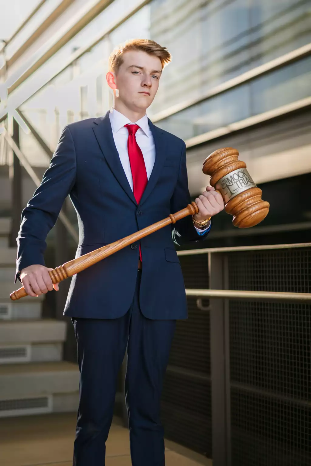 Macon's senior photos taken at Country Square and Downtown Greenville: Young man in a navy suit and red tie stands outside on a staircase, holding an oversized wooden gavel labeled "Mock Trial.".