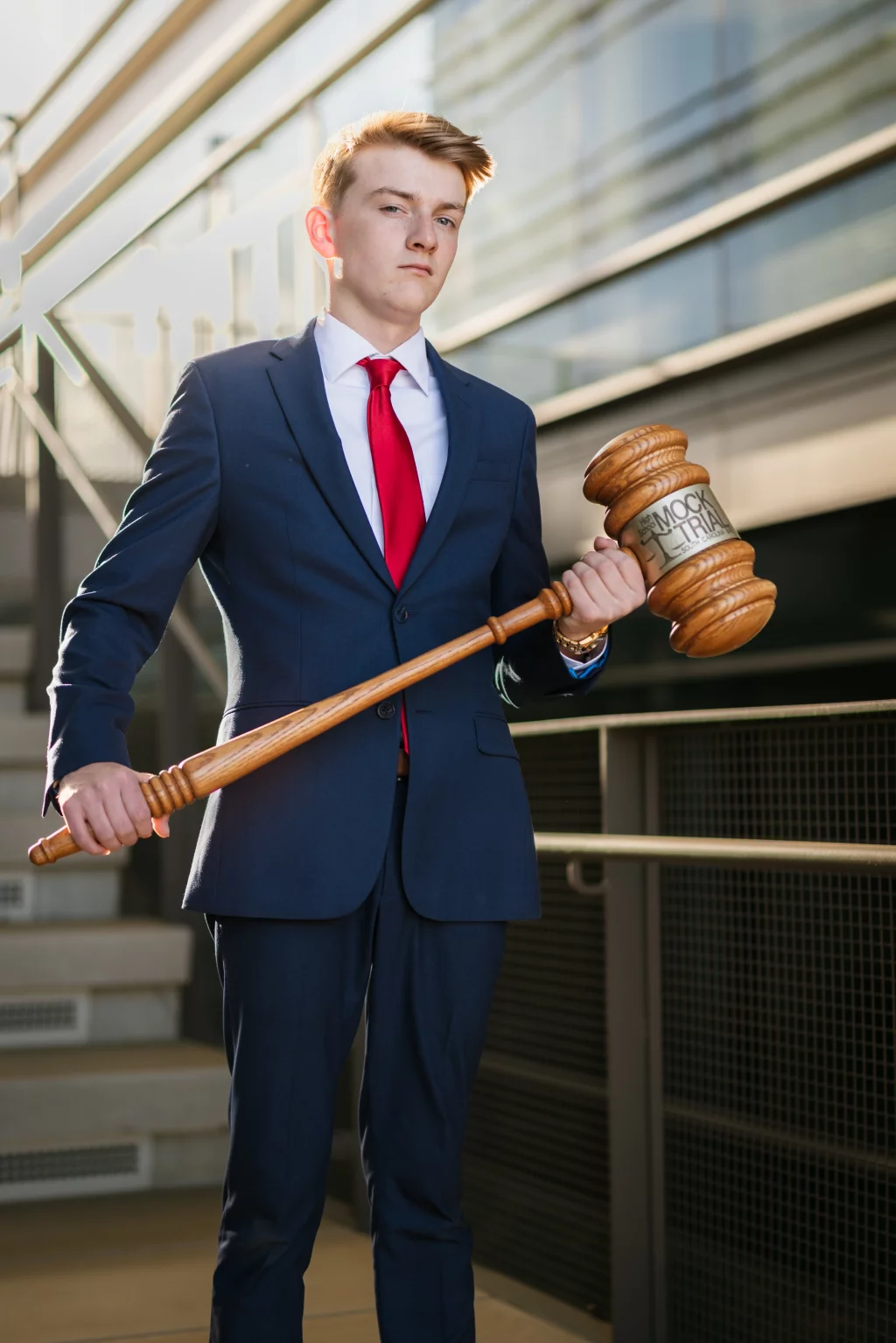 Macon's senior photos taken at Country Square and Downtown Greenville: Young man in a navy suit and red tie stands outside on a staircase, holding an oversized wooden gavel labeled "Mock Trial.".