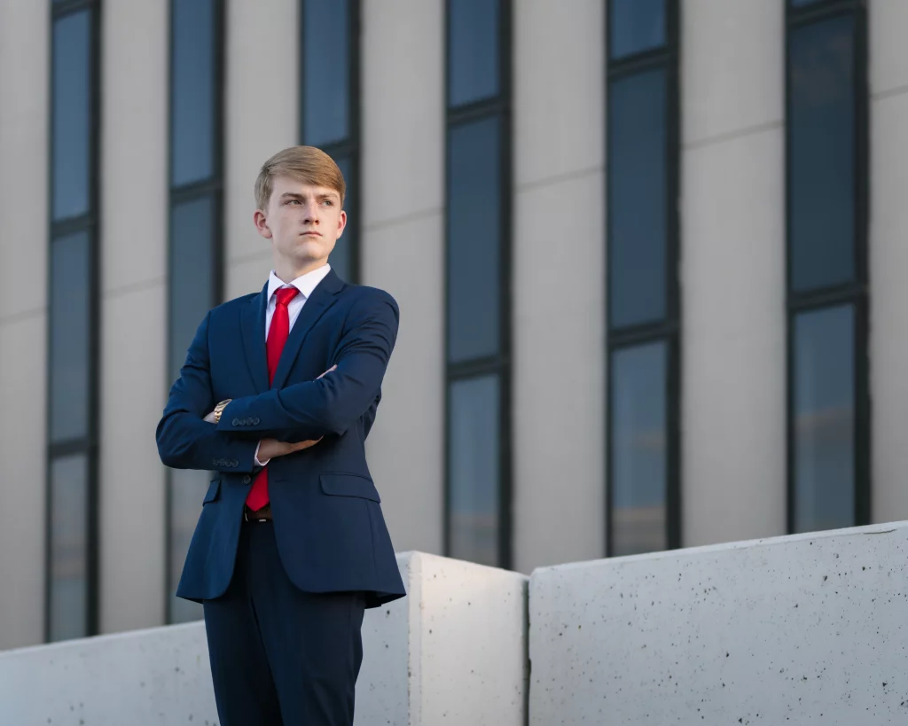 Macon's senior photos taken at County Square and Downtown Greenville: Young man in a navy suit and red tie standing with arms crossed in front of a modern building with tall windows.