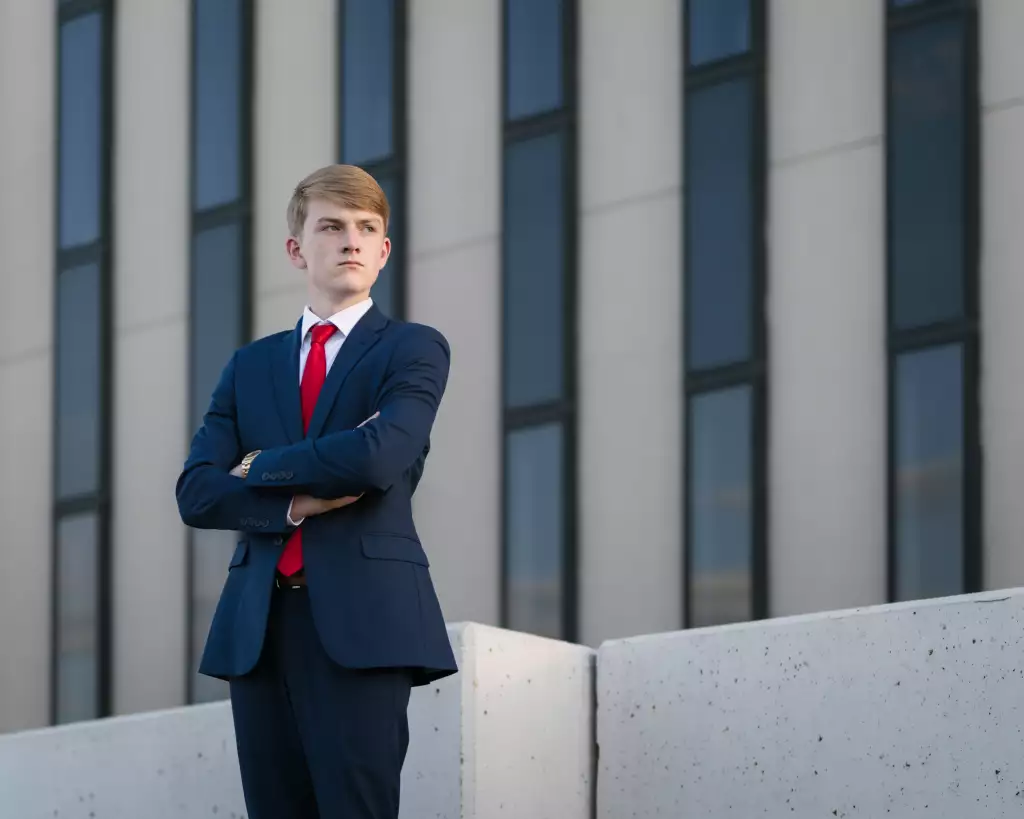 : Young man in a navy suit and red tie standing with arms crossed in front of a modern building with tall windows.