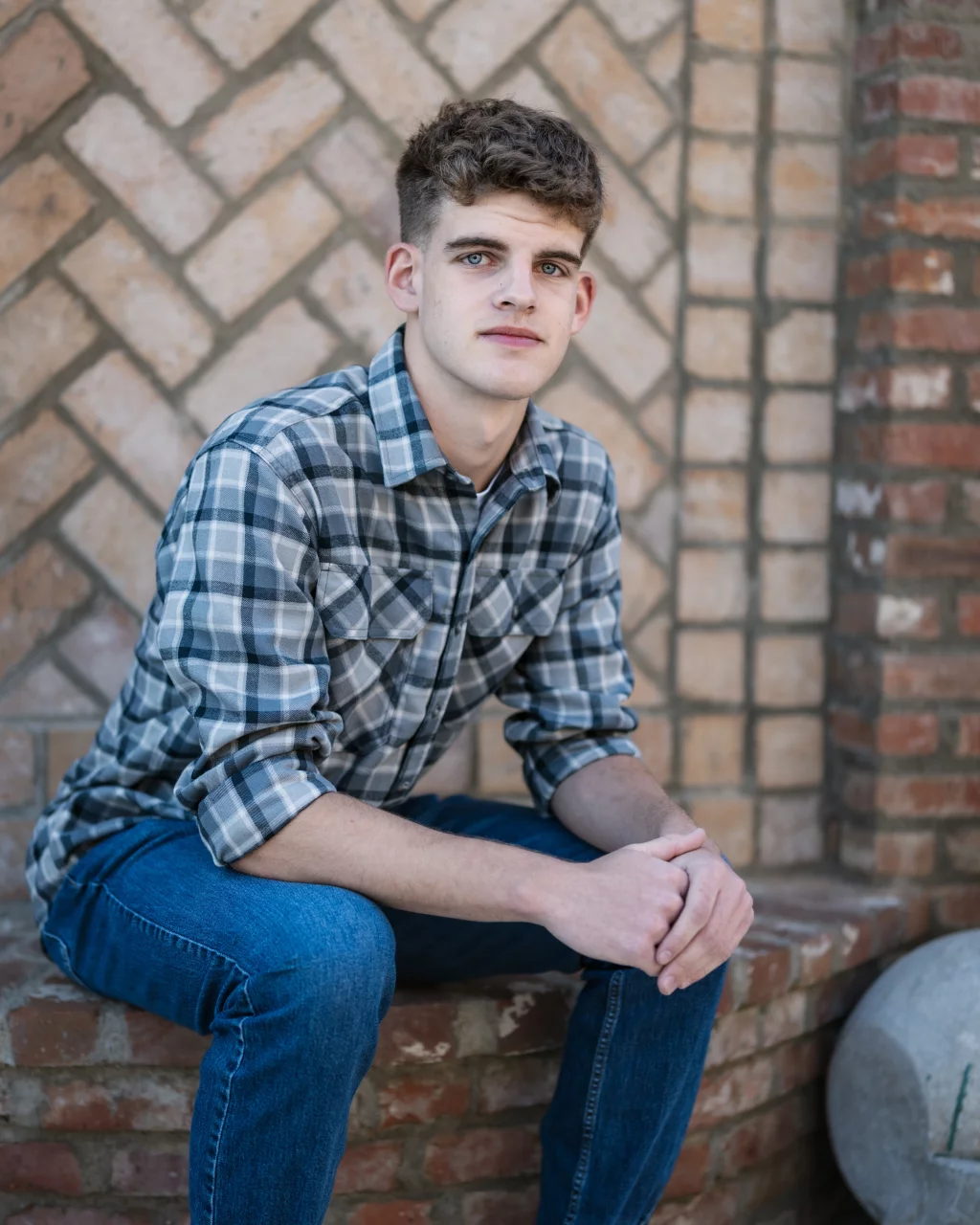 Max's senior photos taken at BridgeWay Station, Simpsonville, SC: Young man with short curly hair wearing a plaid shirt and jeans, sitting on a brick ledge in front of a patterned brick wall, looking at the camera.