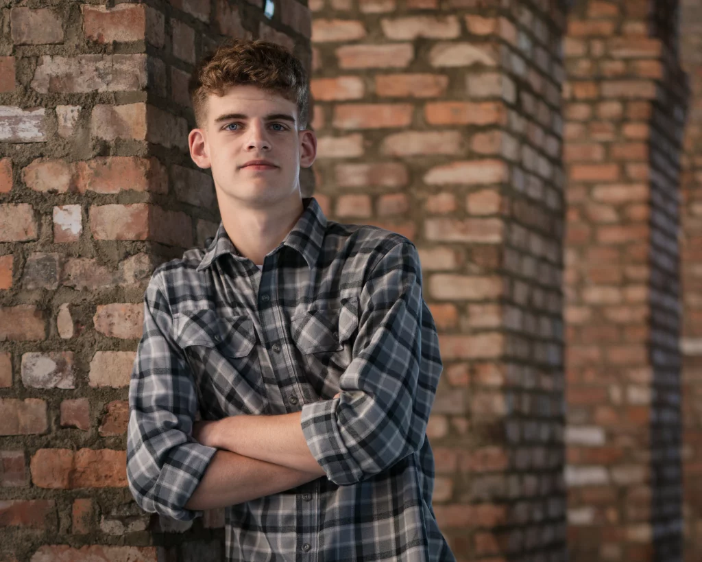 Max's senior photos taken at BridgeWay Station, Simpsonville, SC: Young man with short curly hair and a plaid shirt stands with arms crossed, leaning against a brick pillar in a corridor of similar brick columns.