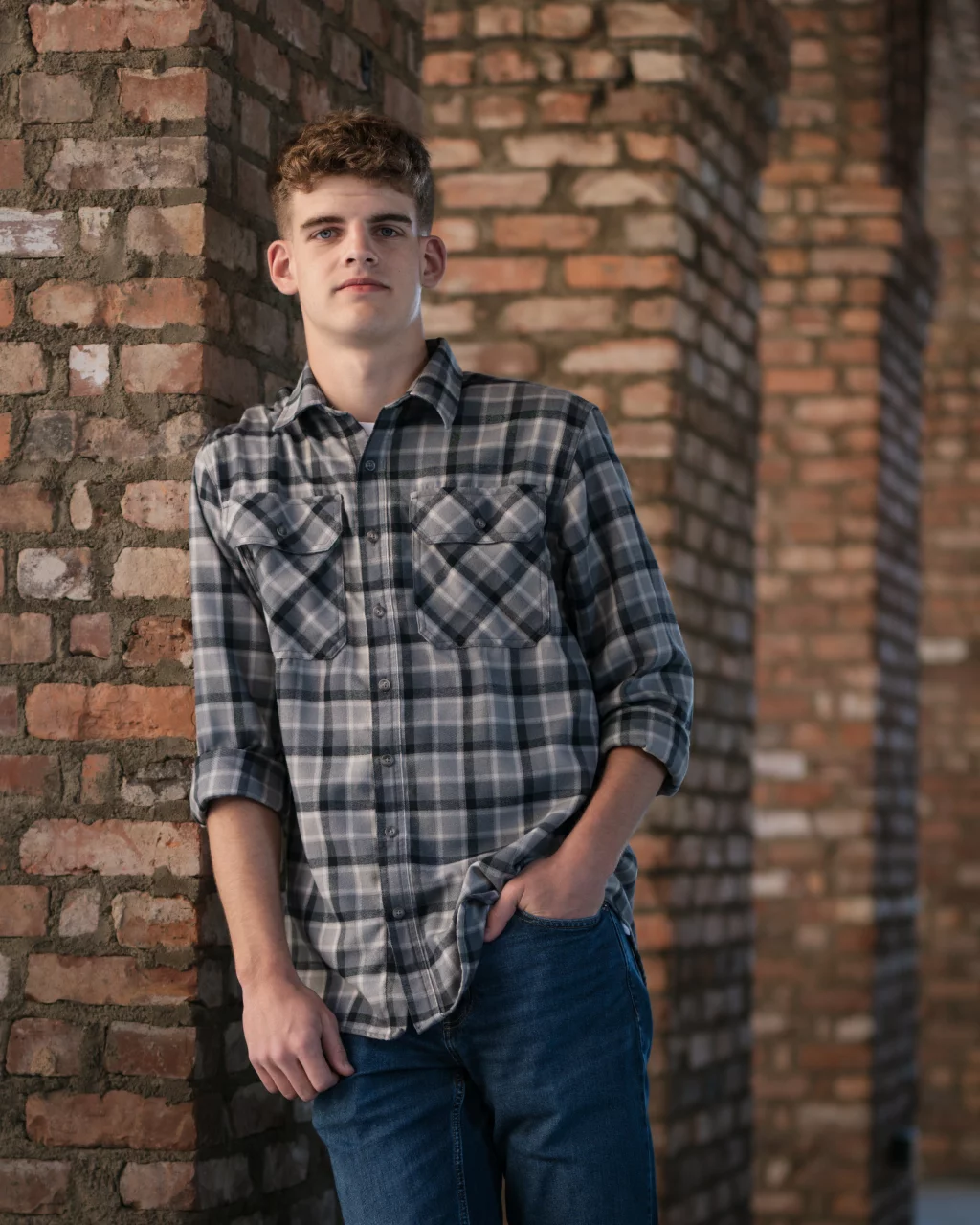Max's senior photos taken at BridgeWay Station, Simpsonville, SC: Young man with short curly hair wearing a gray plaid shirt and blue jeans, leaning against a brick pillar with one hand in his pocket, in a setting with multiple brick arches.