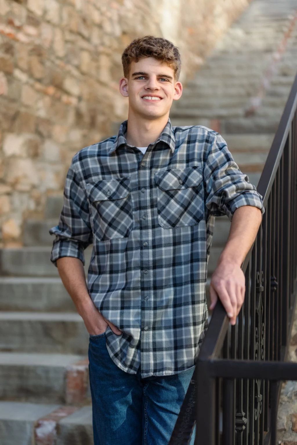 Max's senior photos taken at BridgeWay Station, Simpsonville, SC: Young man with short curly hair wearing a gray plaid shirt and jeans, standing outdoors next to a metal railing with stone steps and a brick wall in the background, smiling at the camera.