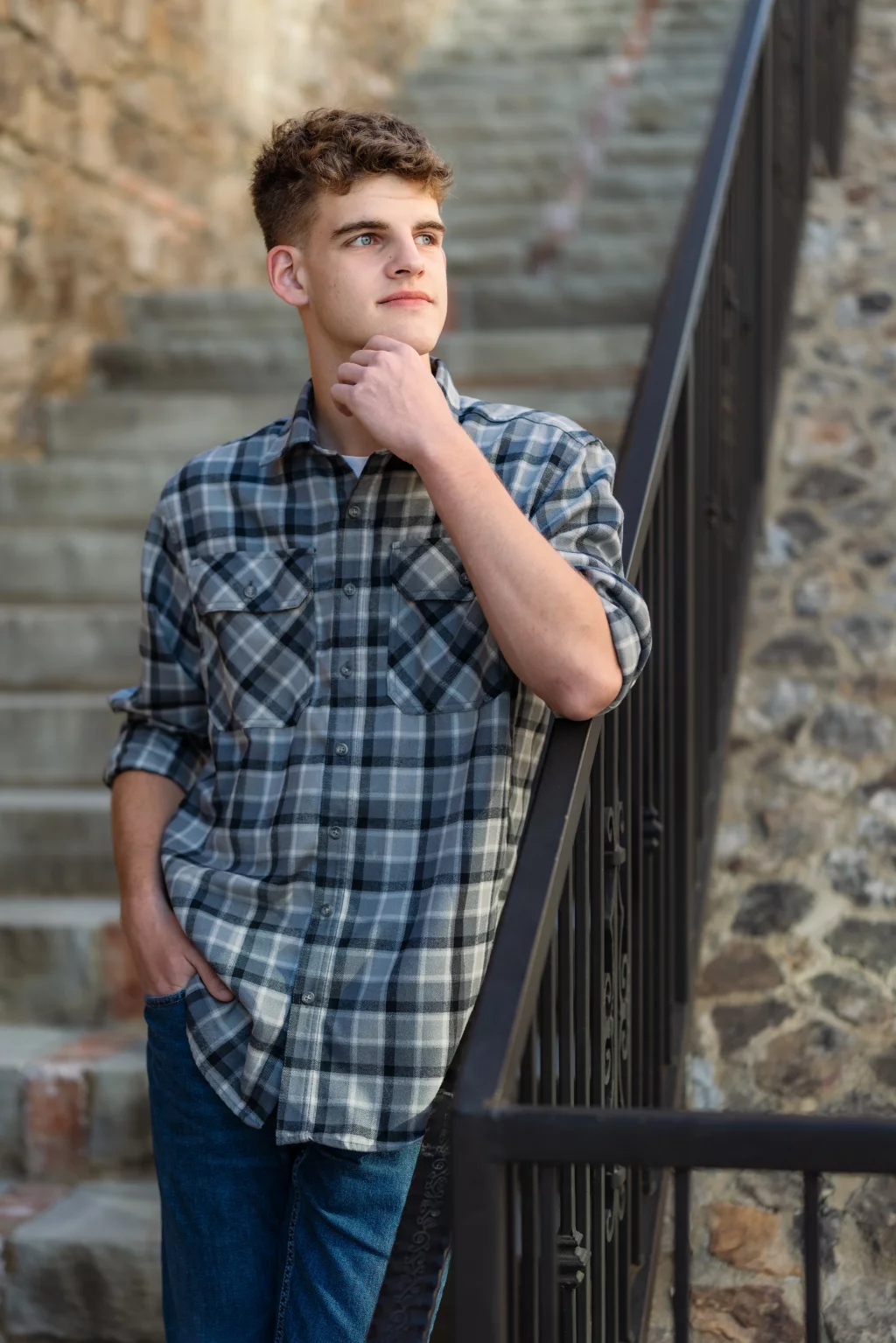 Max's senior photos taken at BridgeWay Station, Simpsonville, SC: Young man in a plaid shirt and jeans standing by a stone staircase and metal railing, looking thoughtfully into the distance with one hand on his chin.