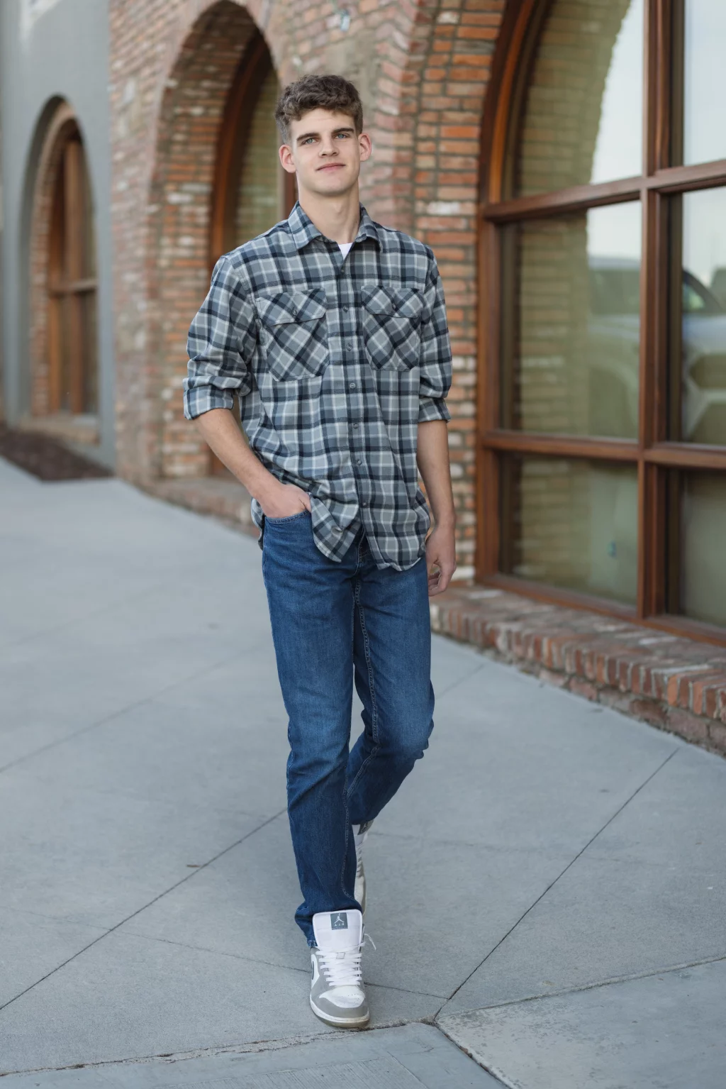 Max's senior photos taken at BridgeWay Station, Simpsonville, SC: Young man with short curly hair wearing a gray plaid shirt, blue jeans, and white sneakers stands on a sidewalk in front of a brick building with large arched windows.