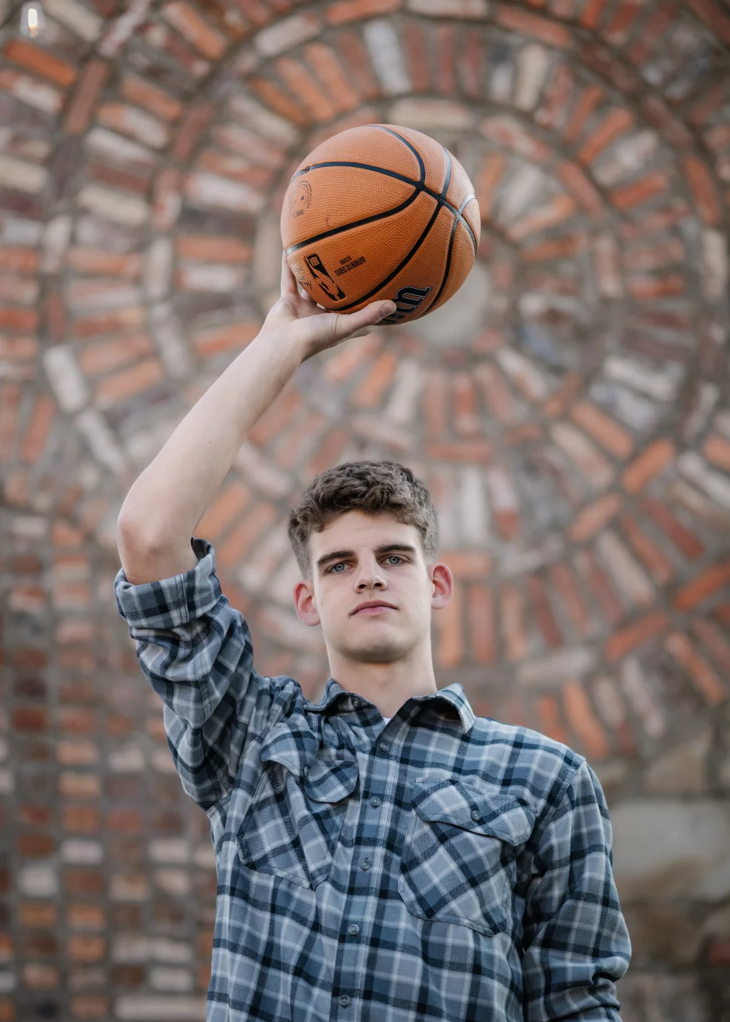 Max's senior photos taken at BridgeWay Station, Simpsonville, SC: Young man in a plaid shirt holding a basketball above his head, standing in front of a circular brick pattern.