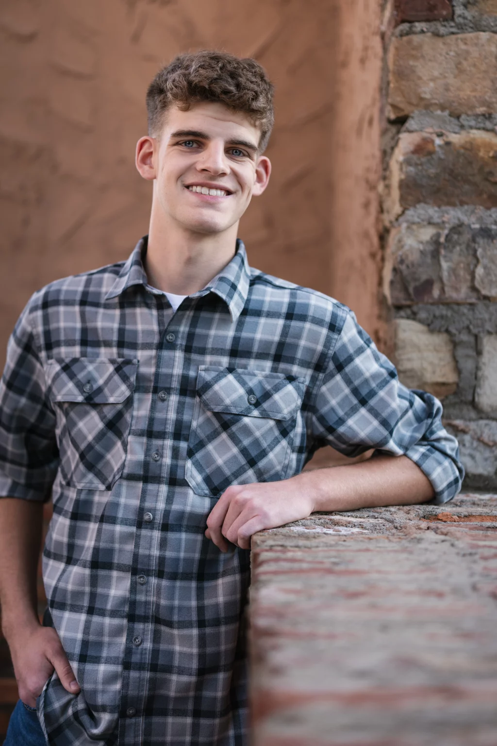 Max's senior photos taken at BridgeWay Station, Simpsonville, SC: Young man with short curly hair wearing a gray plaid shirt, smiling and leaning against a brick wall with one hand in his pocket.