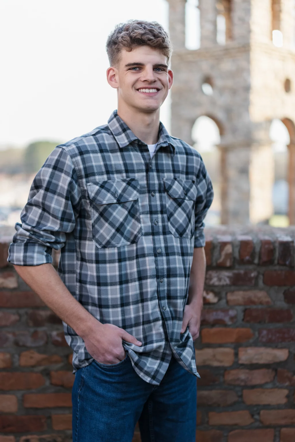 Max's senior photos taken at BridgeWay Station, Simpsonville, SC: Young man in a plaid shirt and jeans standing in front of a brick wall, smiling, with a stone building featuring arches in the background.