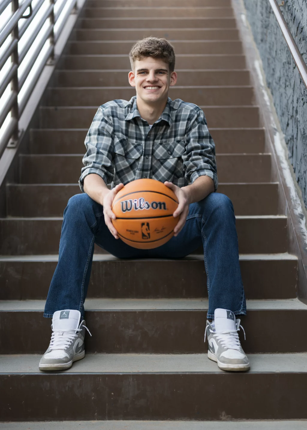 Max's senior photos taken at BridgeWay Station, Simpsonville, SC: Smiling young man in a plaid shirt and jeans sitting on outdoor stairs, holding a basketball.