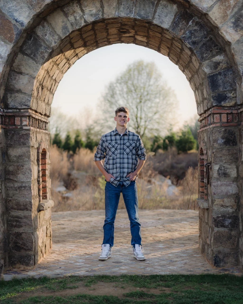 Max's senior photos taken at BridgeWay Station, Simpsonville, SC: Young man in a plaid shirt and jeans standing under a large stone archway outdoors, with trees and greenery in the background.