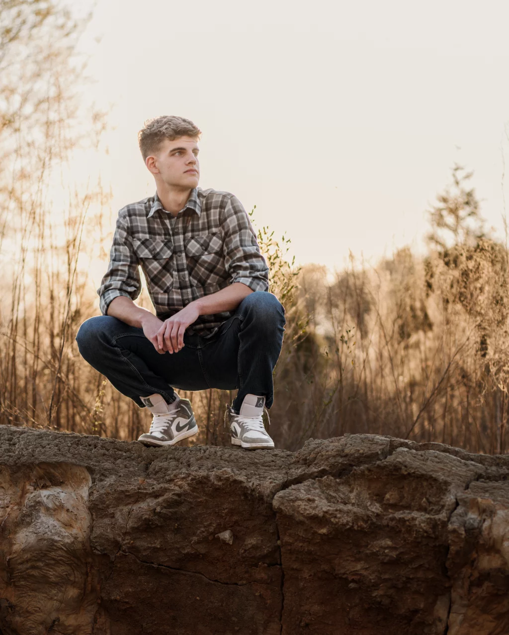 Max's senior photos taken at BridgeWay Station, Simpsonville, SC: Young man in a plaid shirt and jeans squatting on a rocky ledge outdoors, with dry grass and trees in the background at sunset.