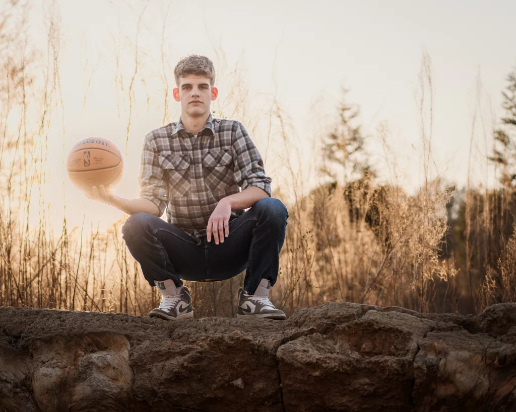 Max's senior photos taken at BridgeWay Station, Simpsonville, SC: Young man in a plaid shirt and jeans squatting on a rock outdoors at sunset, holding a basketball in one hand, with tall dry grass and trees in the background.