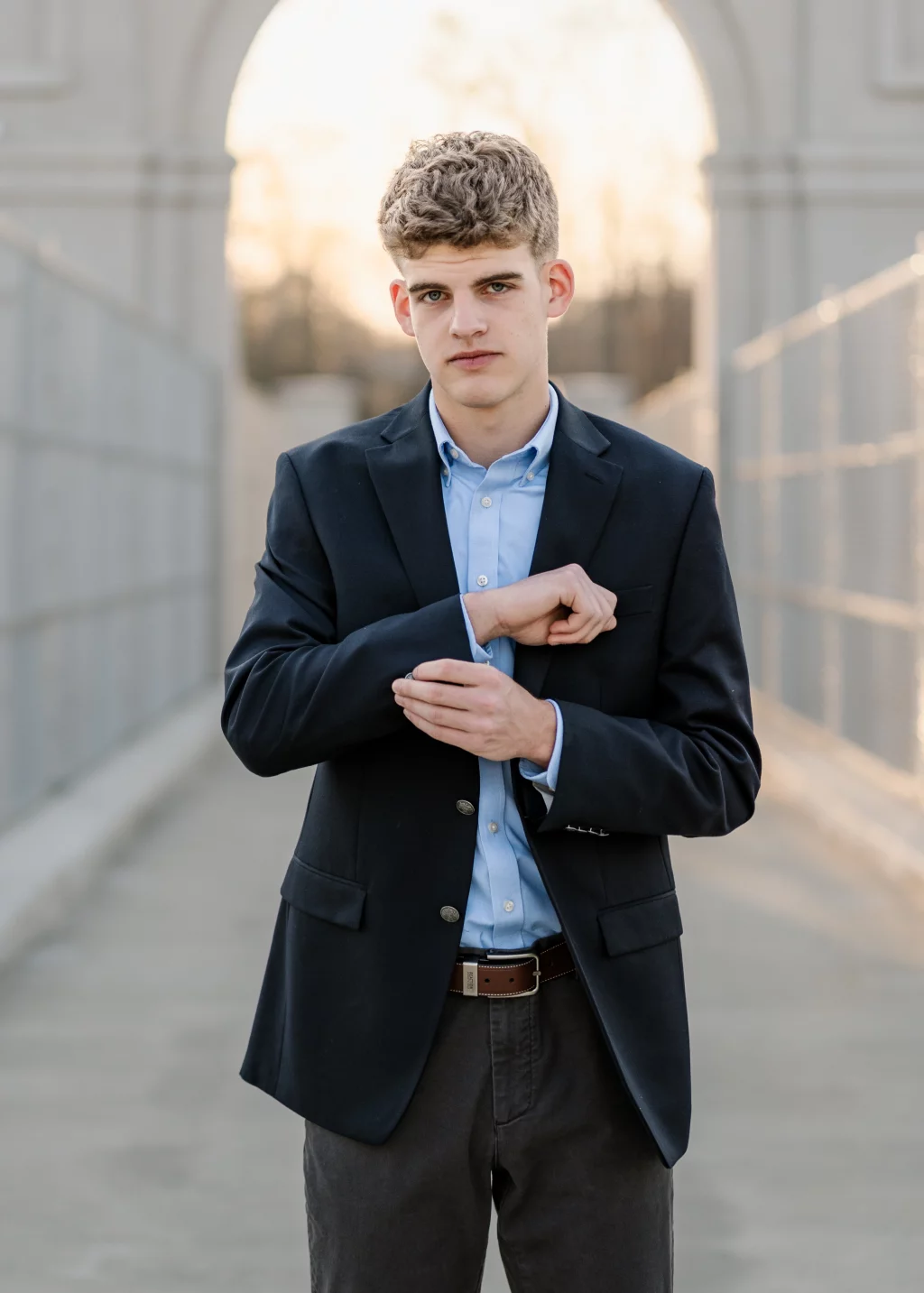 Max's senior photos taken at BridgeWay Station, Simpsonville, SC: Young man with short curly hair wearing a navy blazer and light blue shirt, standing outdoors on a walkway, adjusting his sleeve with an archway and soft sunlight in the background.