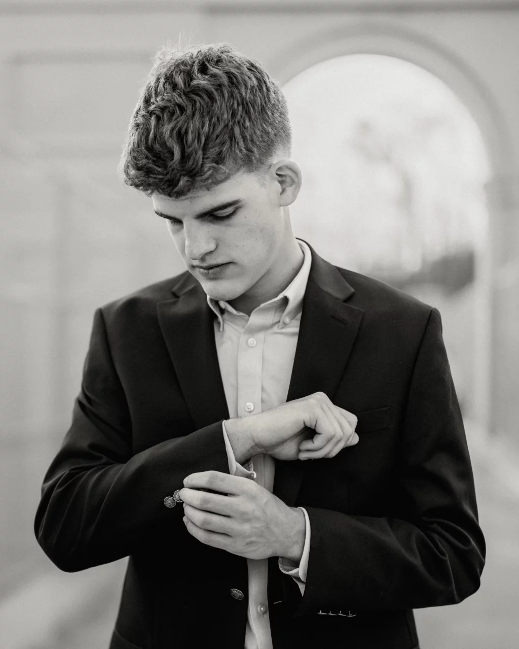 Max's senior photos taken at BridgeWay Station, Simpsonville, SC: Black and white photo of a young man in a suit, looking down and fastening the button on his jacket sleeve, with an arched structure blurred in the background.