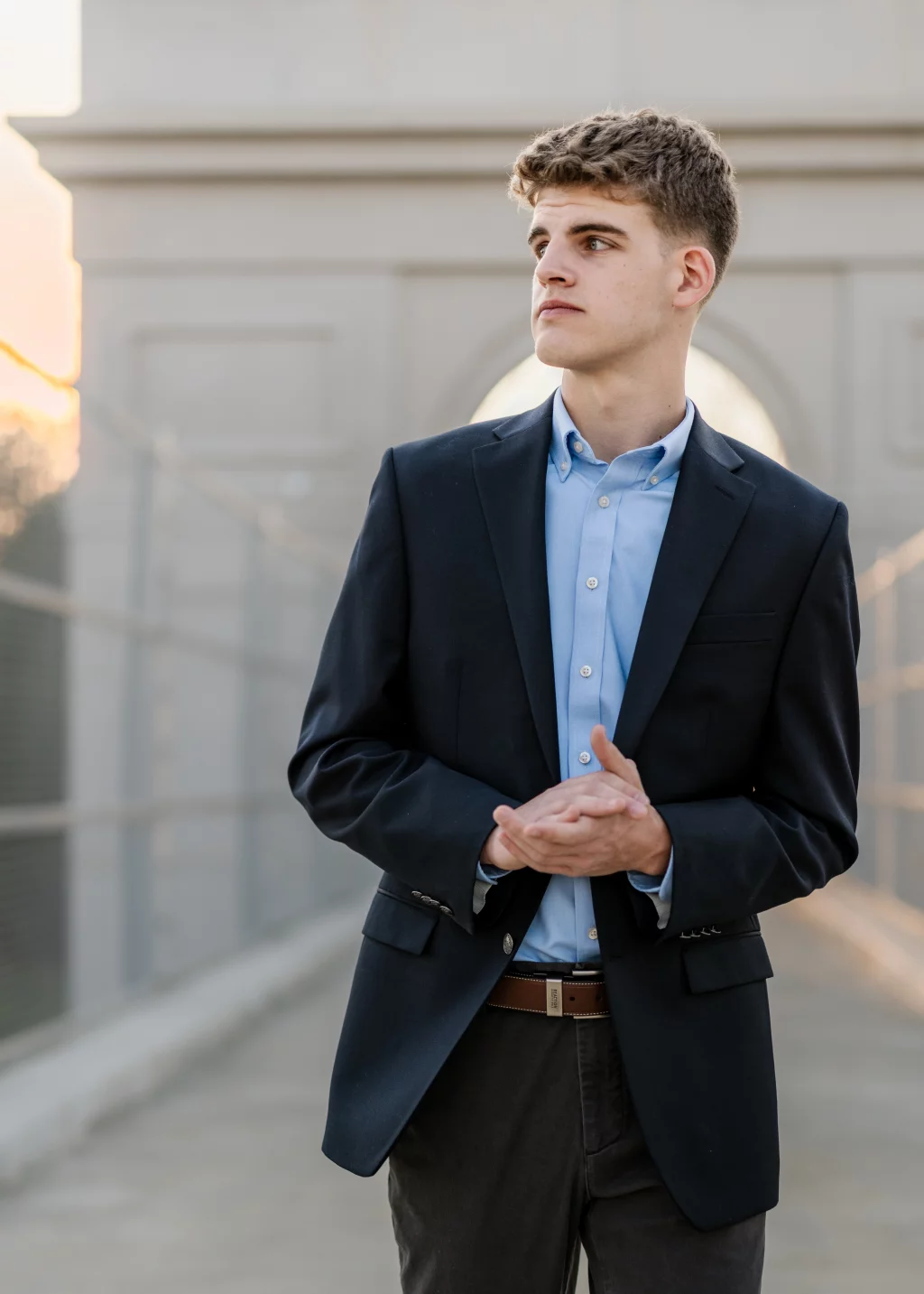 Max's senior photos taken at BridgeWay Station, Simpsonville, SC: Young man in a dark suit jacket and light blue dress shirt standing on a walkway, looking to the side with hands clasped, with an arched architectural structure in the background.