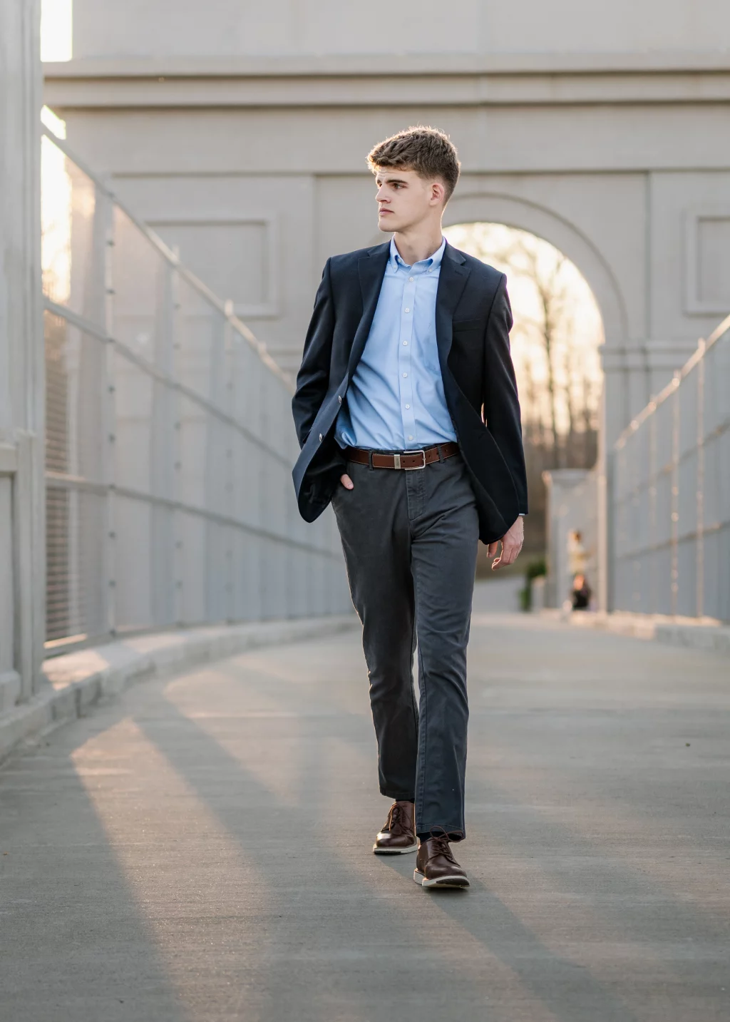 Max's senior photos taken at BridgeWay Station, Simpsonville, SC: Young man in a navy blazer, light blue shirt, and gray pants walking on a bridge with an arched structure in the background, hands in pockets, looking to the side.