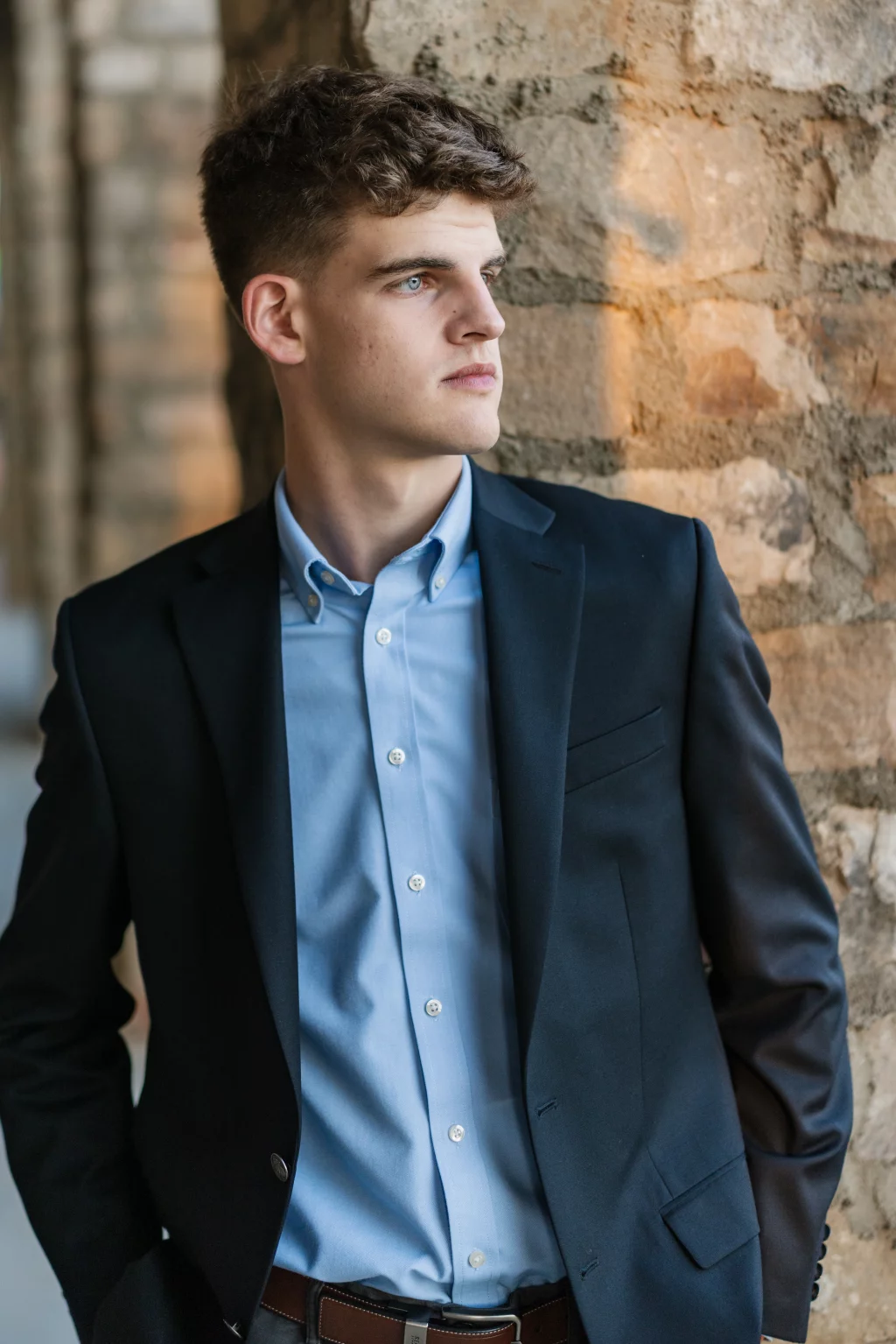 Max's senior photos taken at BridgeWay Station, Simpsonville, SC: Young man in a dark suit jacket and light blue dress shirt standing against a stone wall, looking to the side with a thoughtful expression.