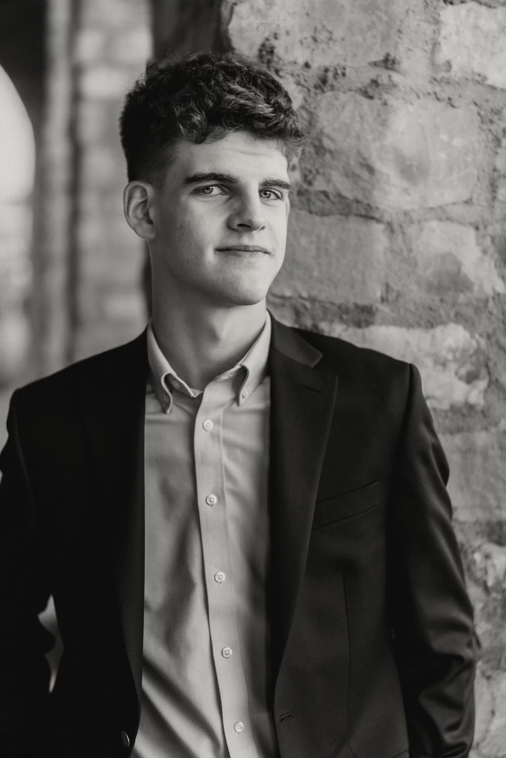 Max's senior photos taken at BridgeWay Station, Simpsonville, SC: Black and white portrait of a young man with short curly hair, wearing a suit jacket and button-up shirt, standing in front of a stone wall.