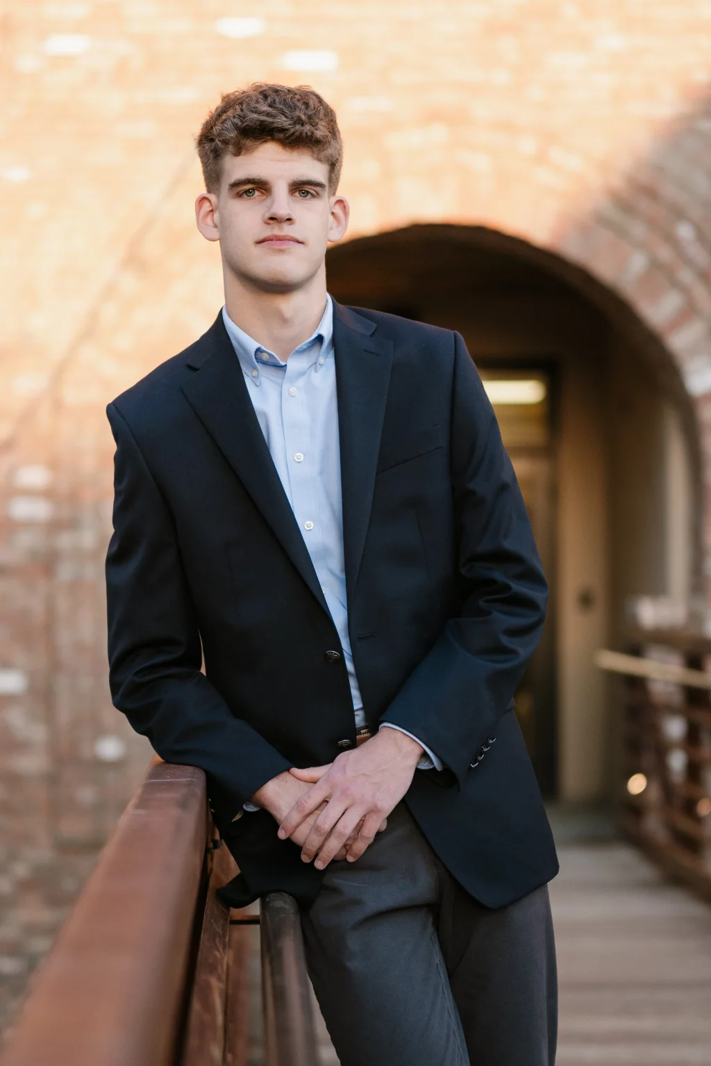 Max's senior photos taken at BridgeWay Station, Simpsonville, SC: Young man in a dark suit jacket and light blue shirt leaning on a wooden railing, standing on a bridge with a brick archway in the background.