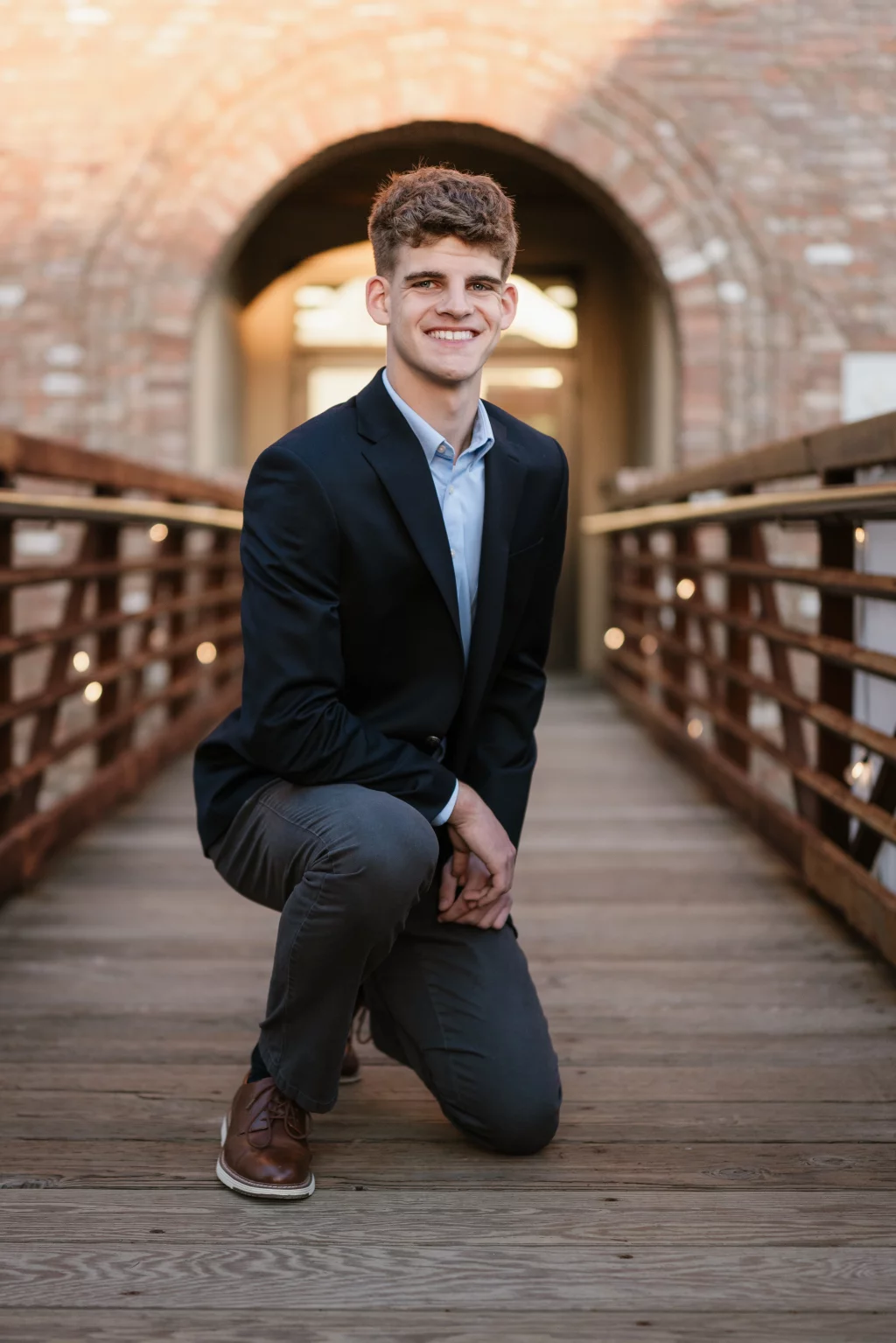 Max's senior photos taken at BridgeWay Station, Simpsonville, SC: Young man in a dark blazer and gray pants kneeling and smiling on a wooden bridge with brick archway in the background.