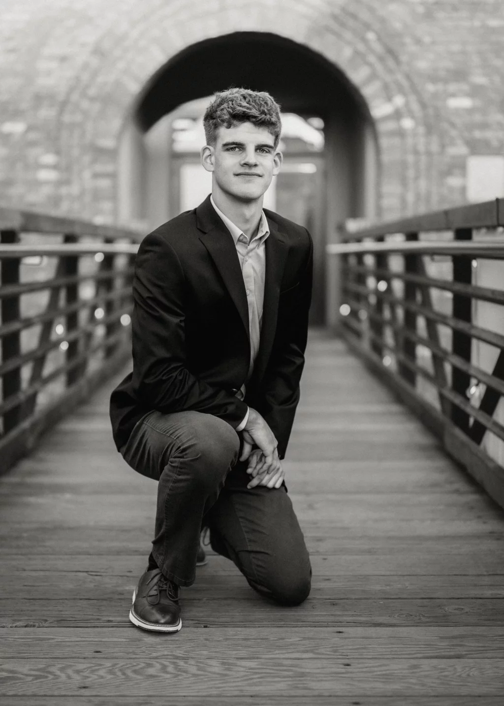 Max's senior photos taken at BridgeWay Station, Simpsonville, SC: Black and white photo of a young man in a suit jacket kneeling on a wooden bridge, with a brick archway in the background.
