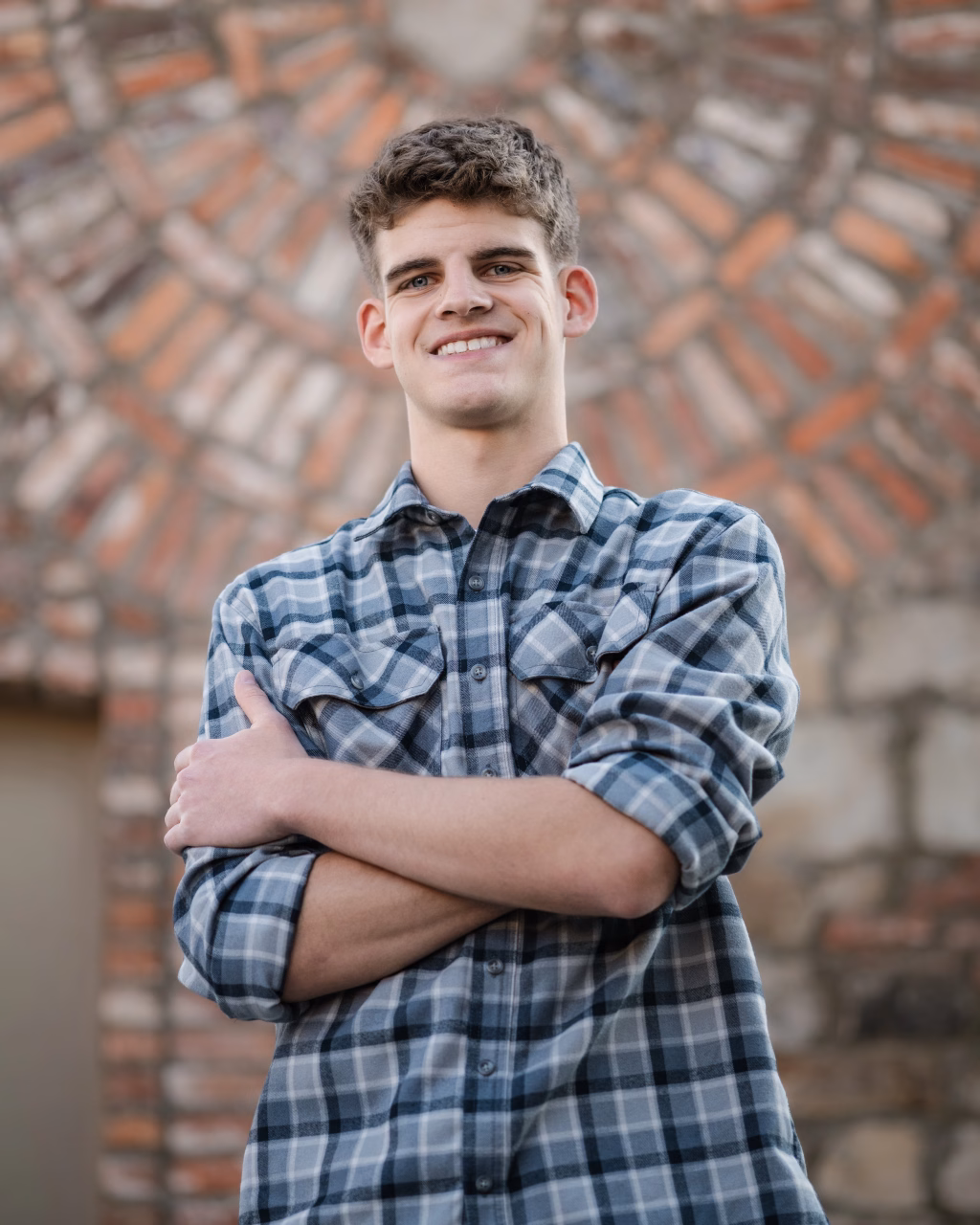 Max's senior photos taken at BridgeWay Station, Simpsonville, SC: Young man in a blue plaid shirt standing with arms crossed, smiling in front of a patterned brick and stone wall.