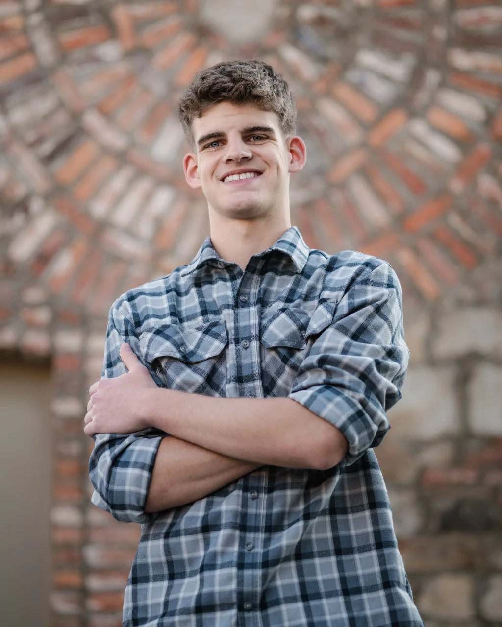 Max's senior photos taken at BridgeWay Station, Simpsonville, SC: Young man in a blue plaid shirt standing with arms crossed, smiling in front of a patterned brick and stone wall.