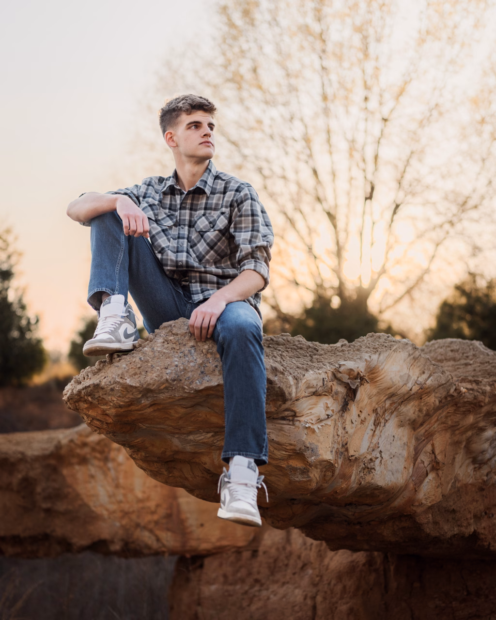 Max's senior photos taken at BridgeWay Station, Simpsonville, SC: Young man in a plaid shirt and jeans sits on a large rock ledge outdoors at sunset, looking off to the side; bare trees and soft sunlight fill the background.