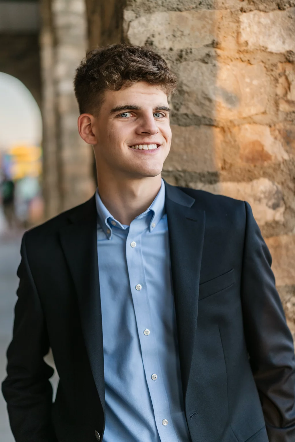 Max's senior photos taken at BridgeWay Station, Simpsonville, SC: Young man with short curly hair wearing a dark suit jacket and light blue dress shirt, smiling and leaning against a stone wall.