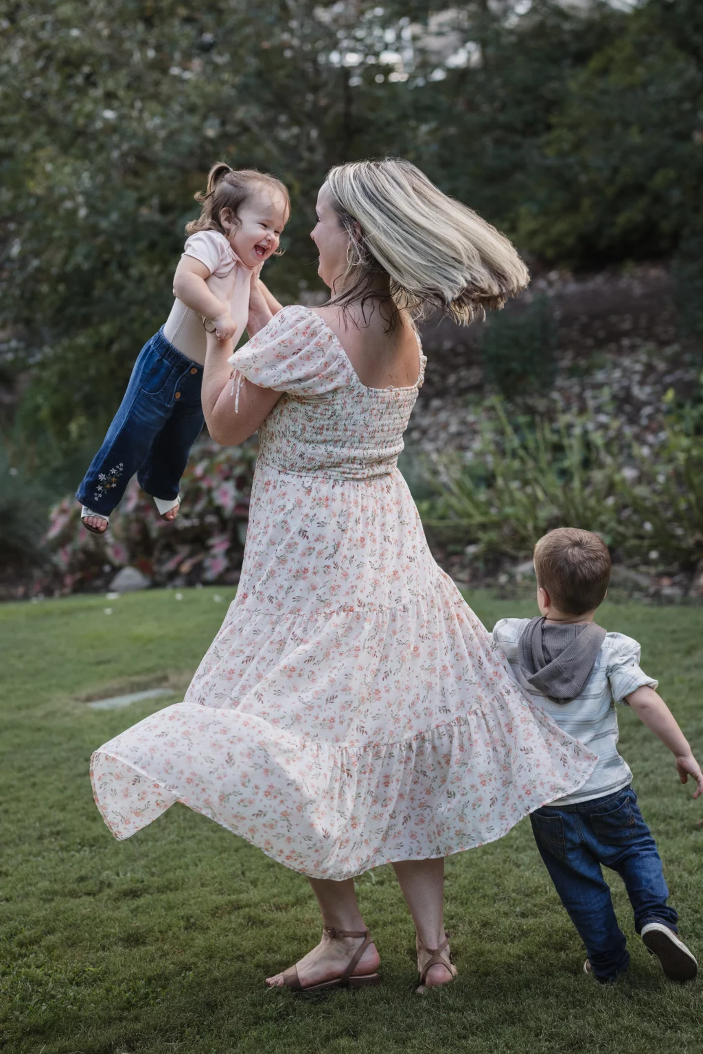 Family session at the Rock Quarry Garden.: A woman in a floral dress playfully lifts a laughing toddler while another young child stands beside her in a grassy garden.